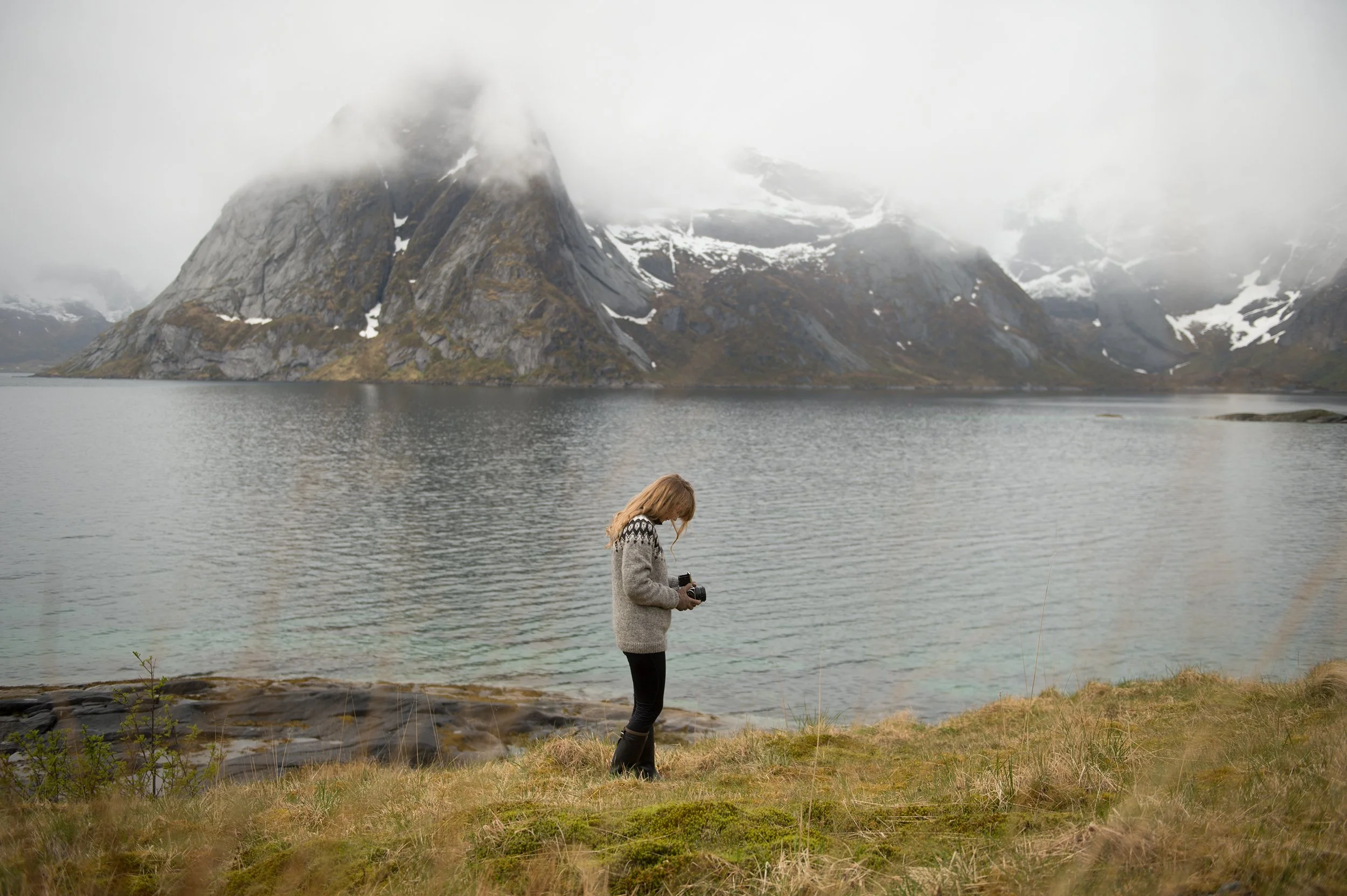 woman in a grey sweater stands next to a norwegian fjord  with foggy mountains in the distance