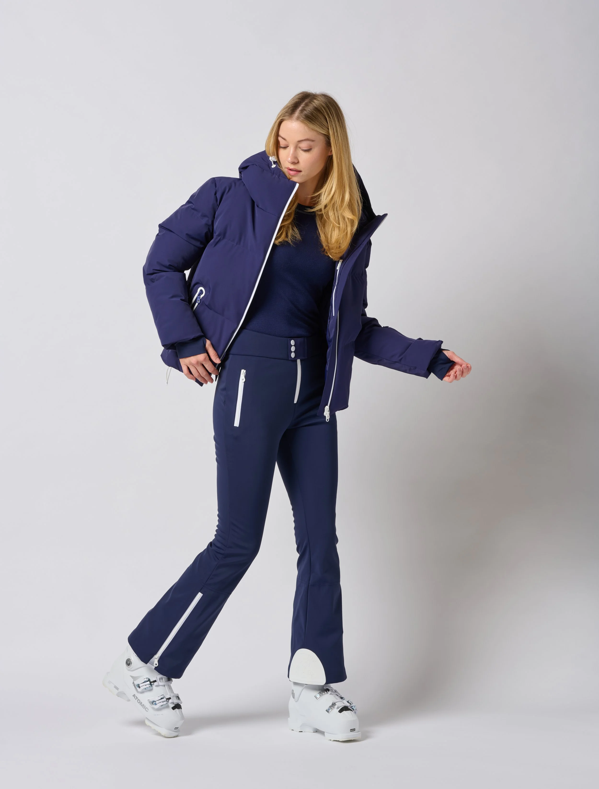 A woman in a navy blue Jorde Skiwear winter jacket, matching pants, and white ski boots, standing against a plain light gray background in a Burlington Vermont photography studio.
