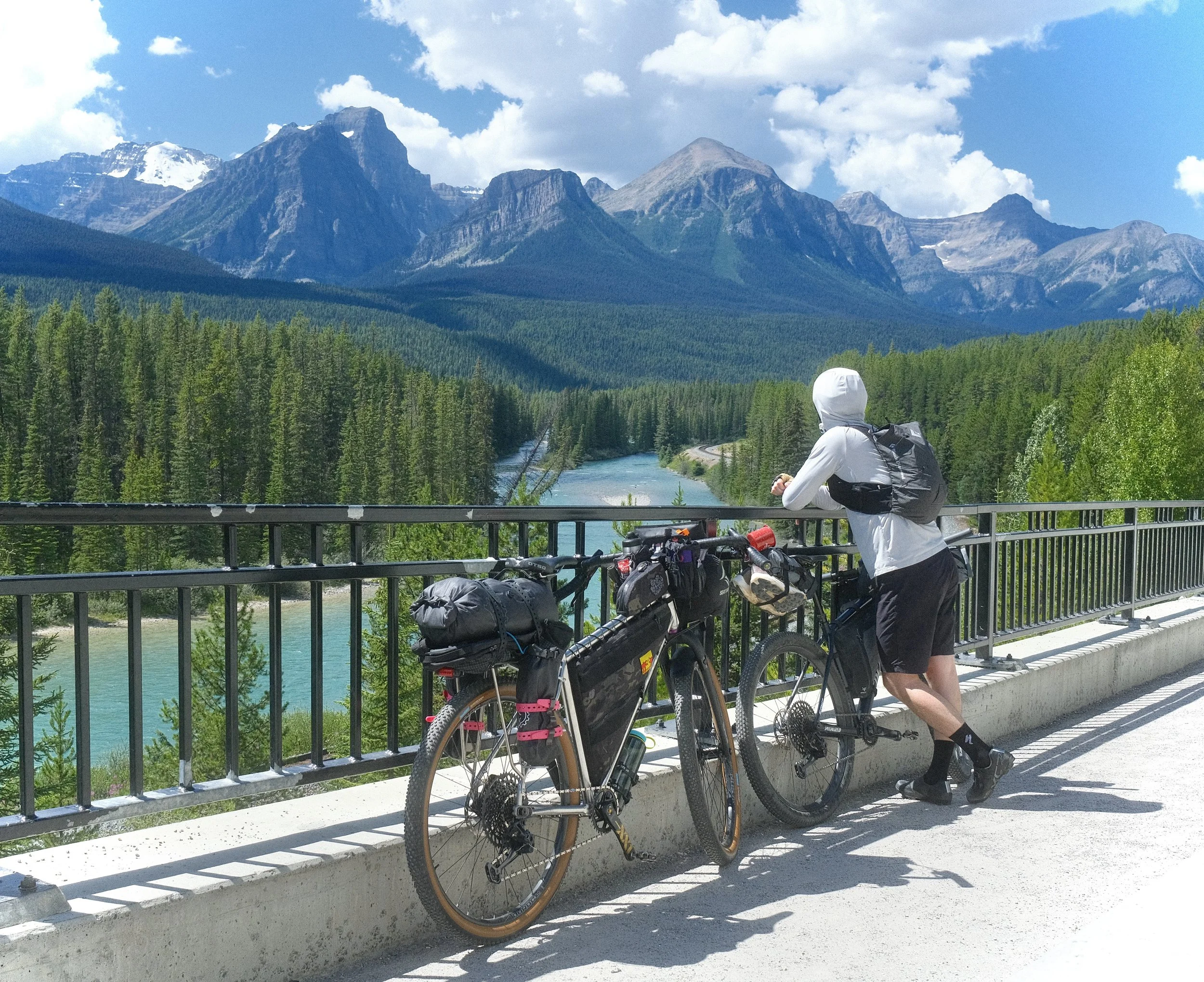 A person with a bikepacking bike leaning on a railing, looking at the scenic Bow River and dense forest with mountains in the background under a partly cloudy sky in Banff National Park