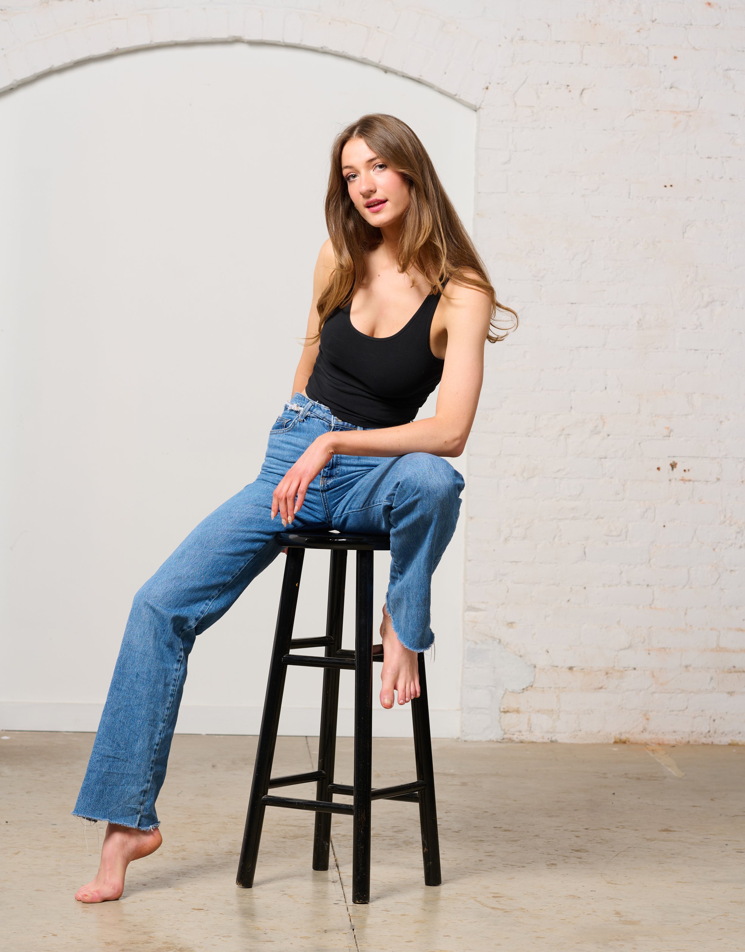 Woman sitting on a black stool, wearing a black tank top and blue jeans, against a white brick wall for a studio fashion shoot in Burlington Vermont.