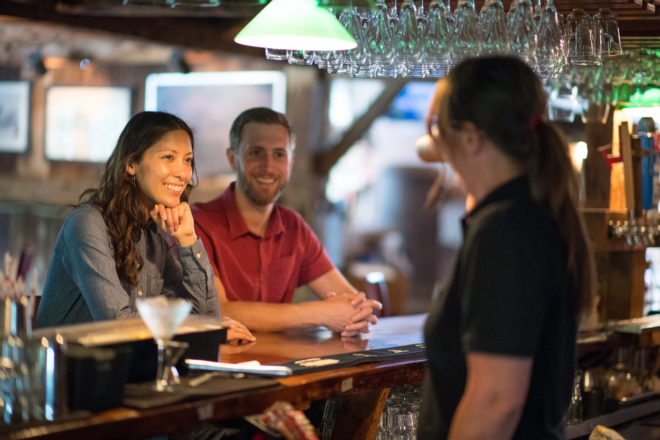 man and woman on a date sit at a bar while talking to the bartender
