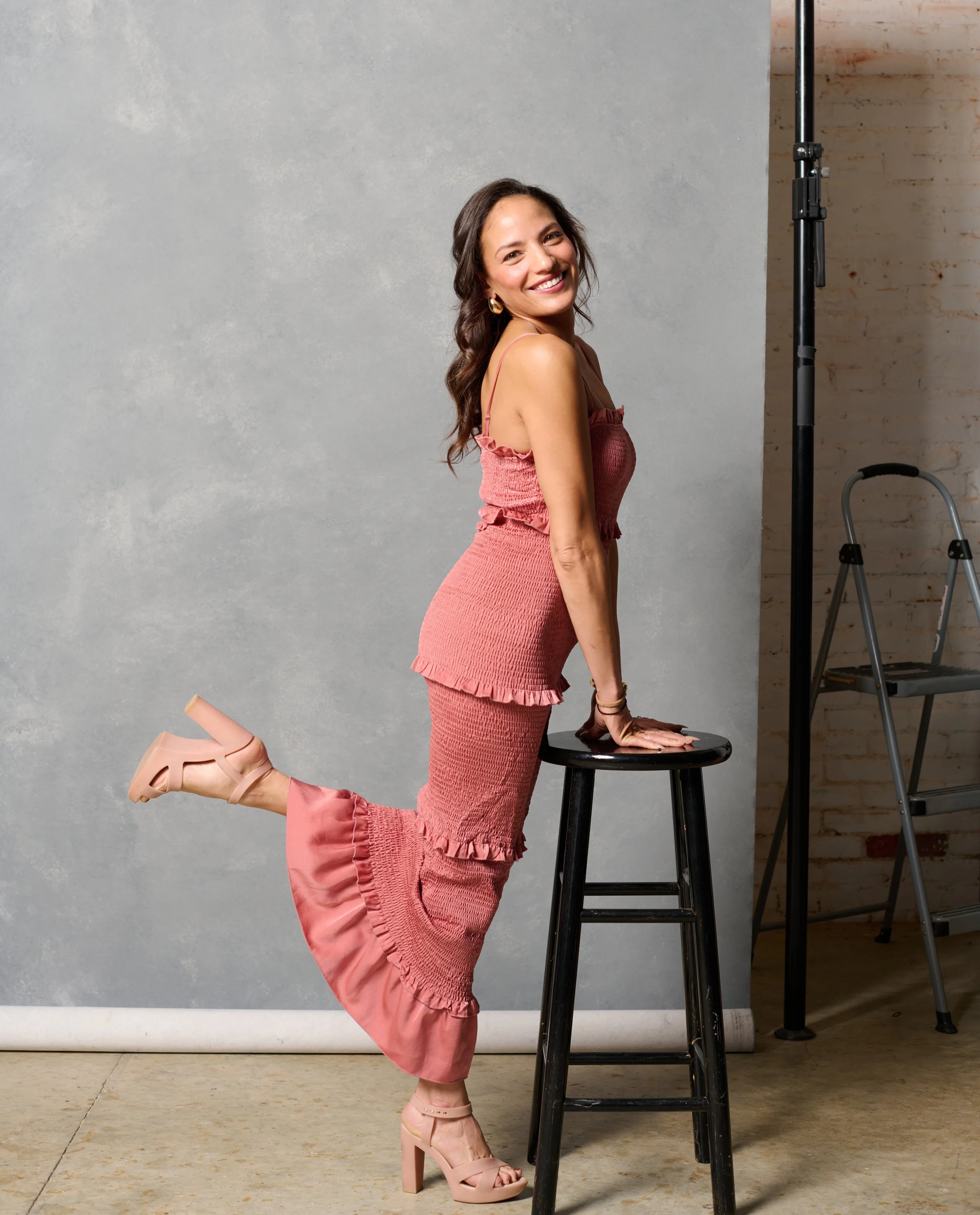 a dark haired woman leans on a stool and smiles at camera during a photo shoot