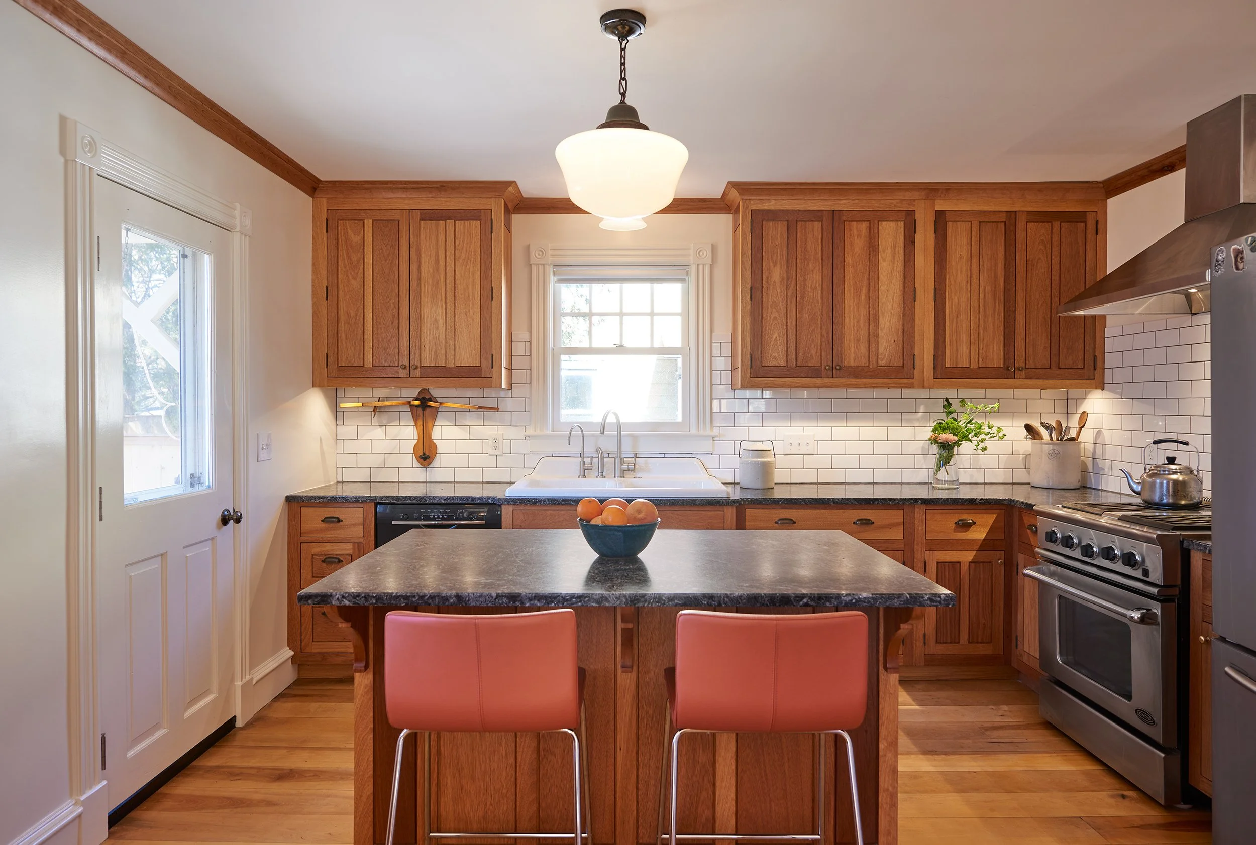 Warm kitchen with wooden cabinets, a central island with black countertop, two orange chairs, stainless steel appliances, white subway tiles, and a bowl of oranges.