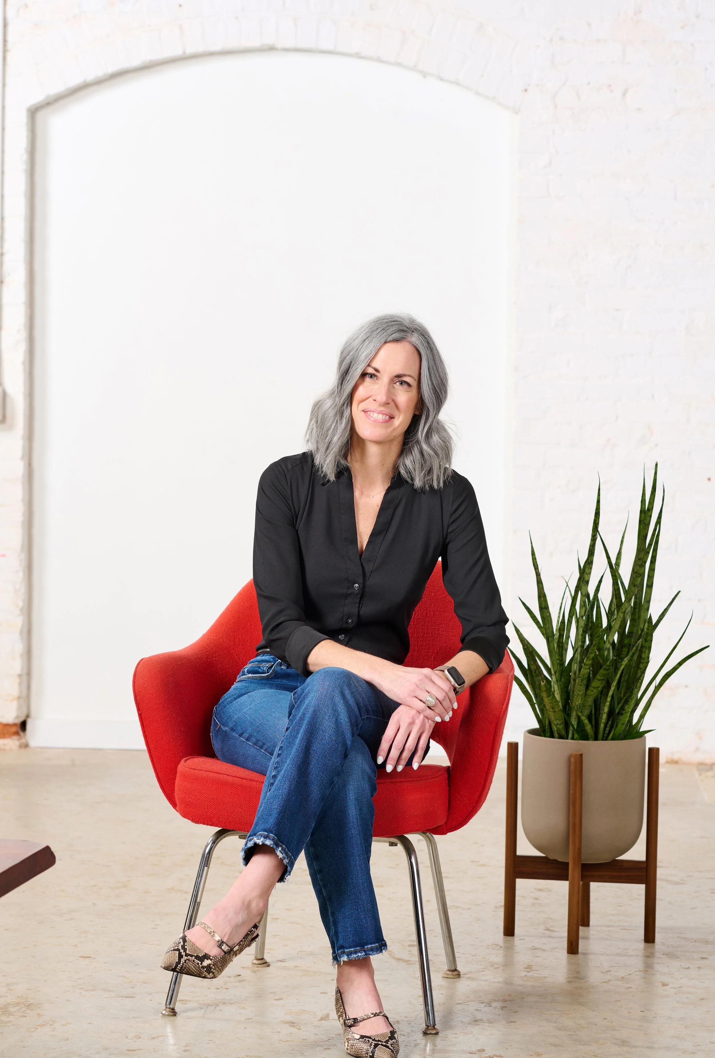 Meg McKeen sits in a red chair with green plant next to her while wearing a black shirt and blue jeans