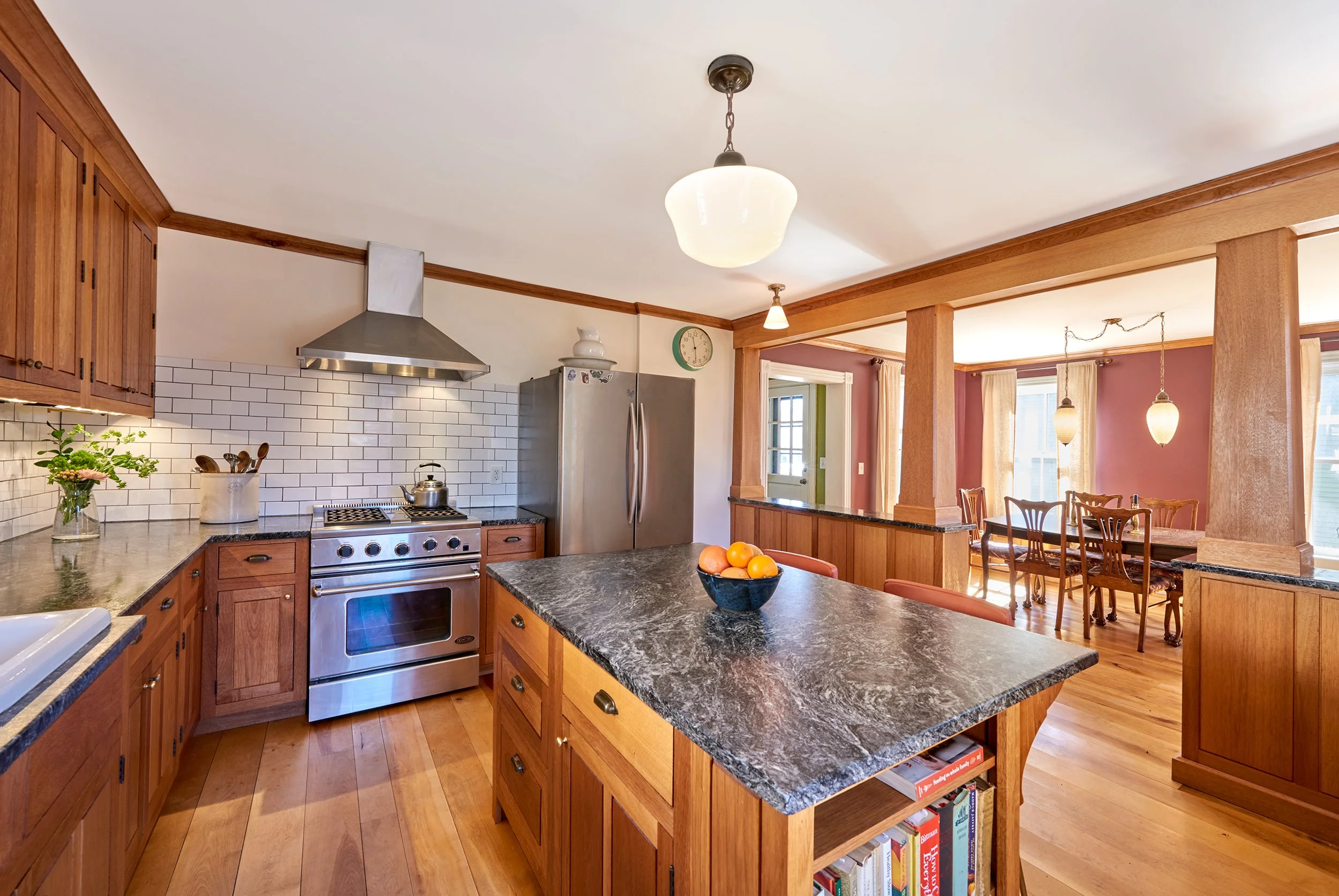 Spacious kitchen with wooden cabinets, stainless steel stove and fridge, black granite countertops, and a central island with a bowl of oranges. The kitchen opens into a dining area with a wooden table and chairs, and pendant lighting.