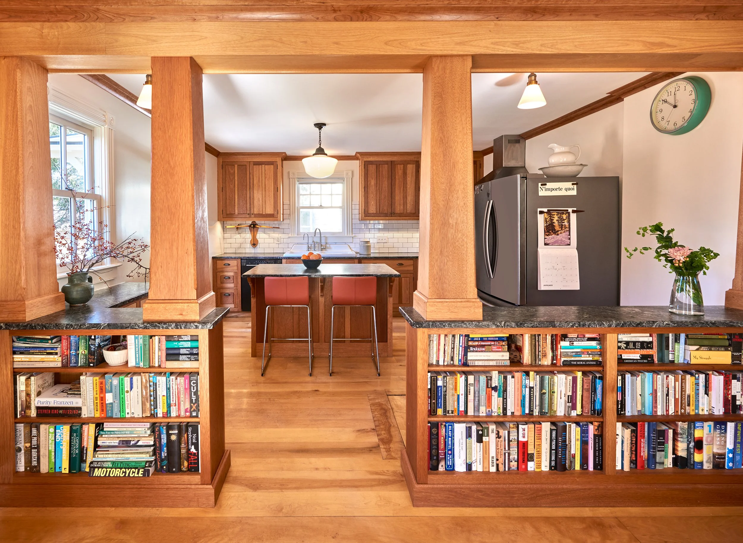 Kitchen with wooden cabinets, a large refrigerator, kitchen island, bookshelves, and a clock on the wall.