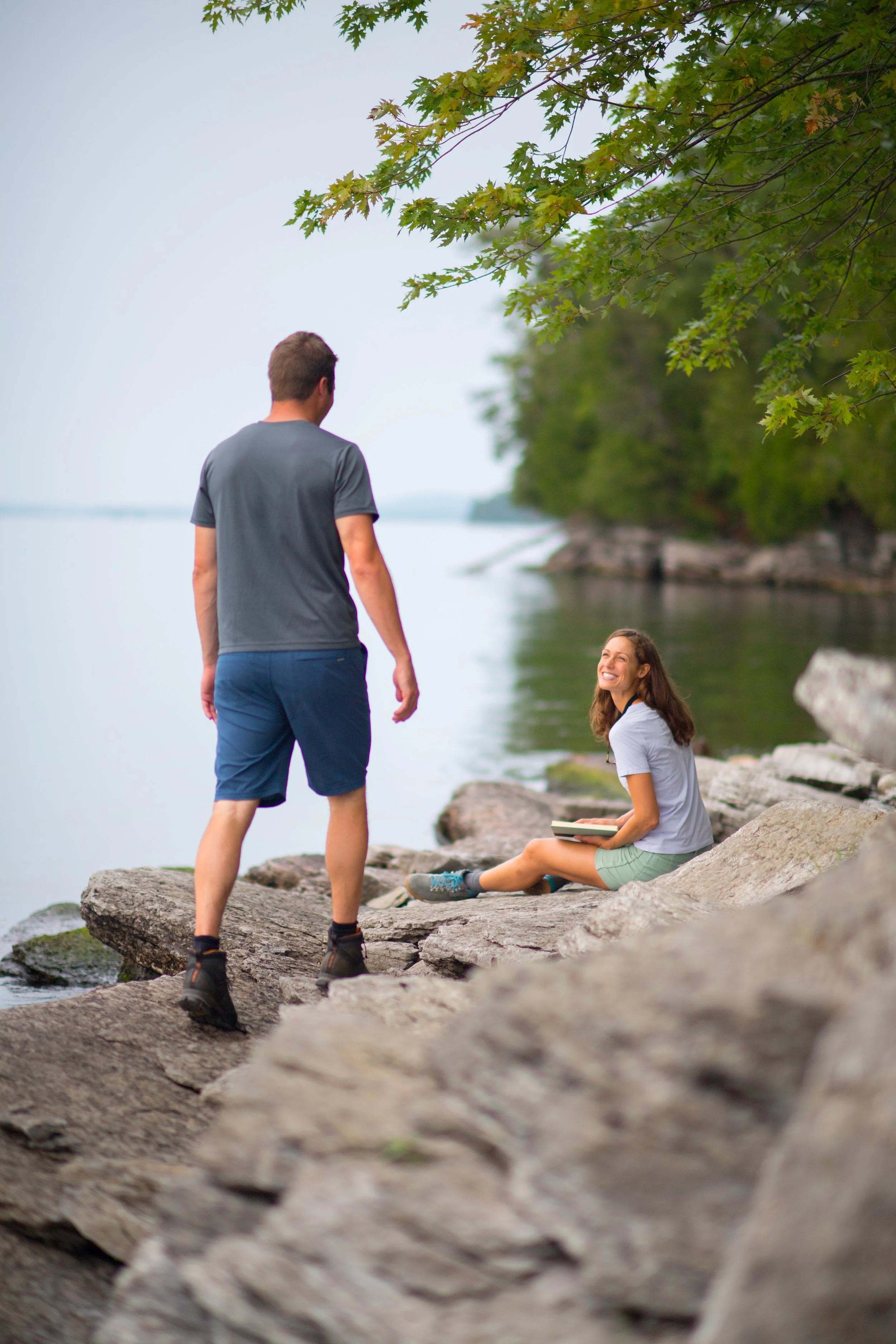 lifestyle photography of a man walking up to his girlfriend as she sits on a rocky shore in vermont while reading a book