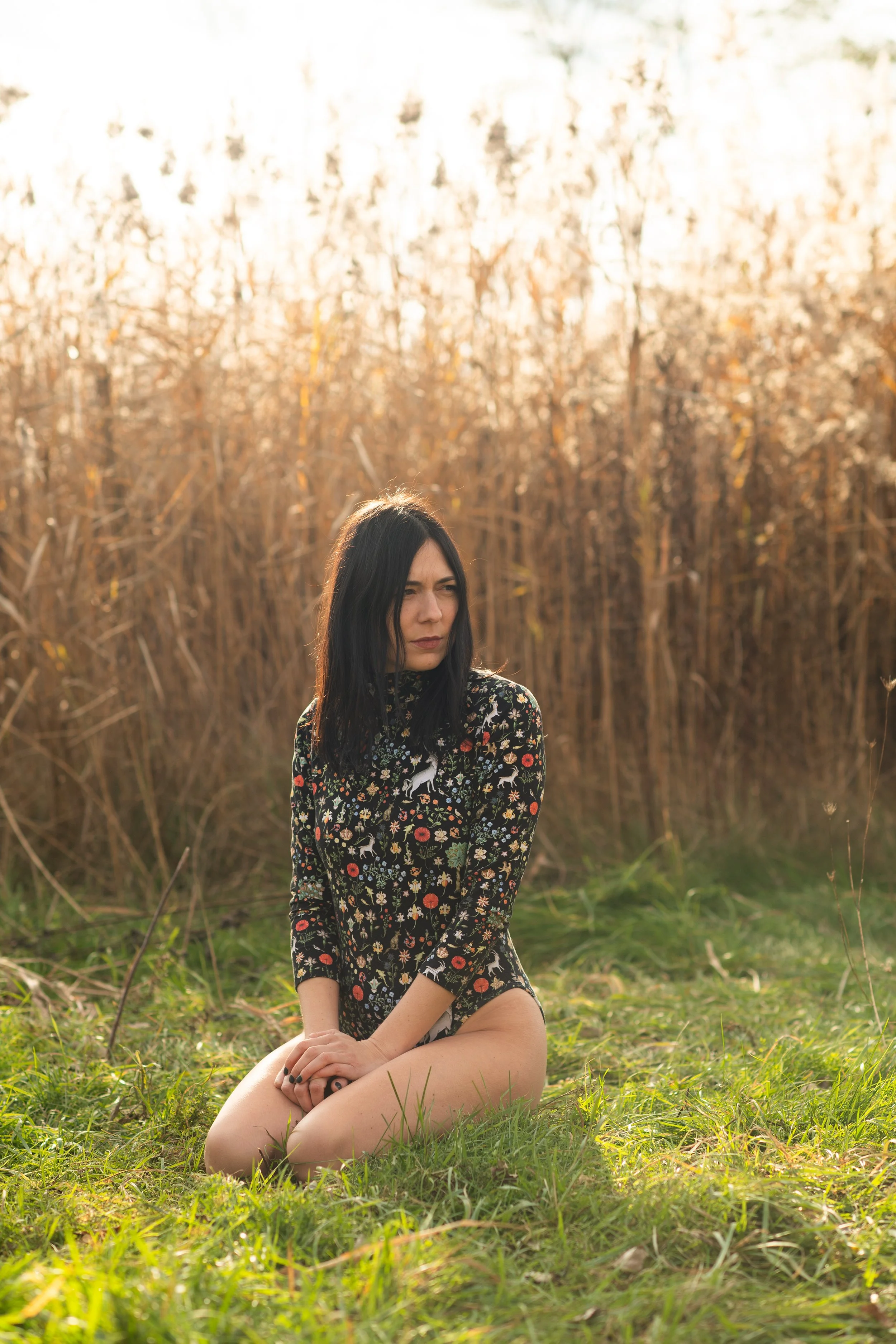 outdoor fashion photography photo shoot of woman kneeling in grass with reeds and sunset  behind her