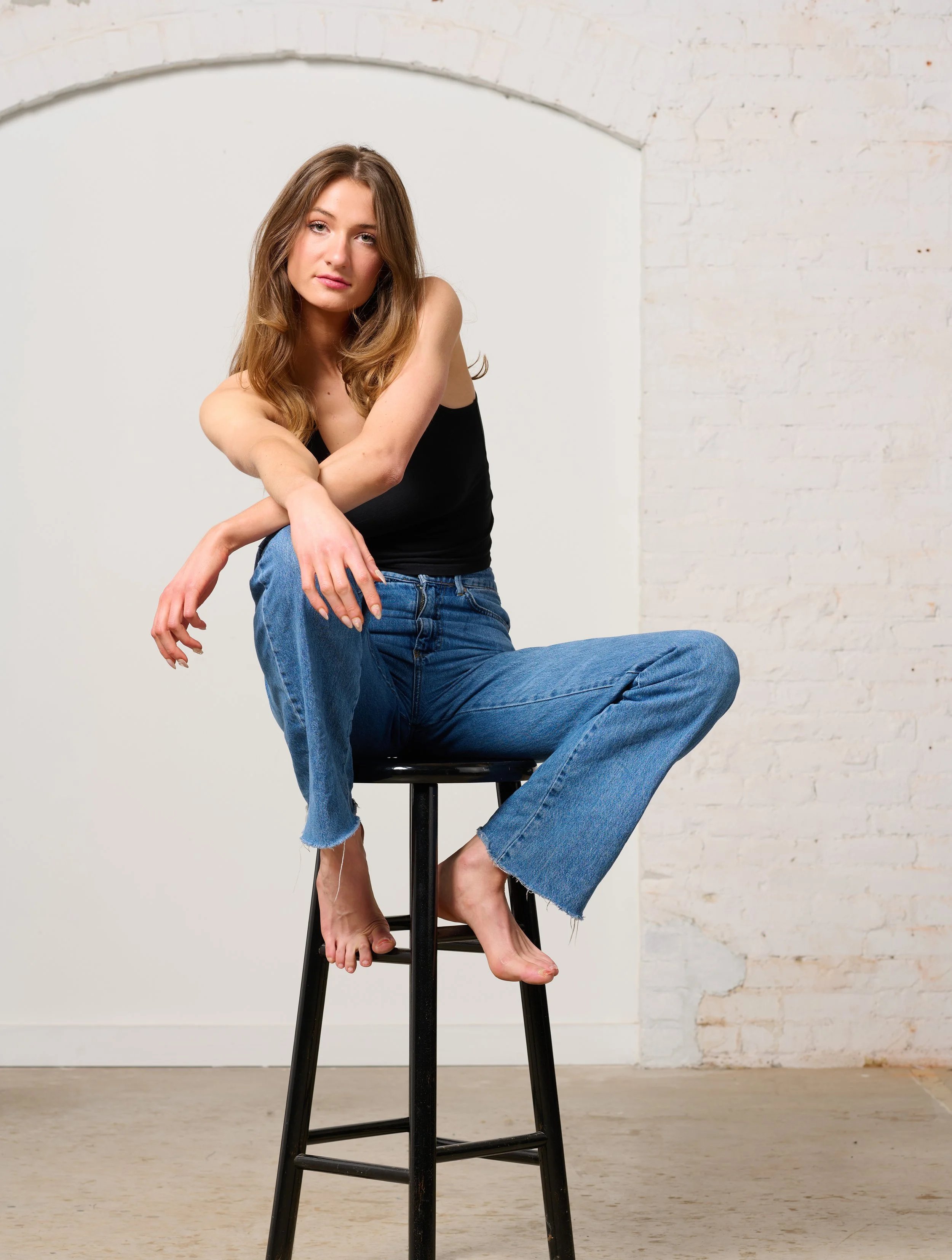 A model sitting on a stool posing for a burlington vermont studio fashion shoot, wearing a black tank top and blue jeans, against a white brick wall background in a Burlington Vermont photography studio.