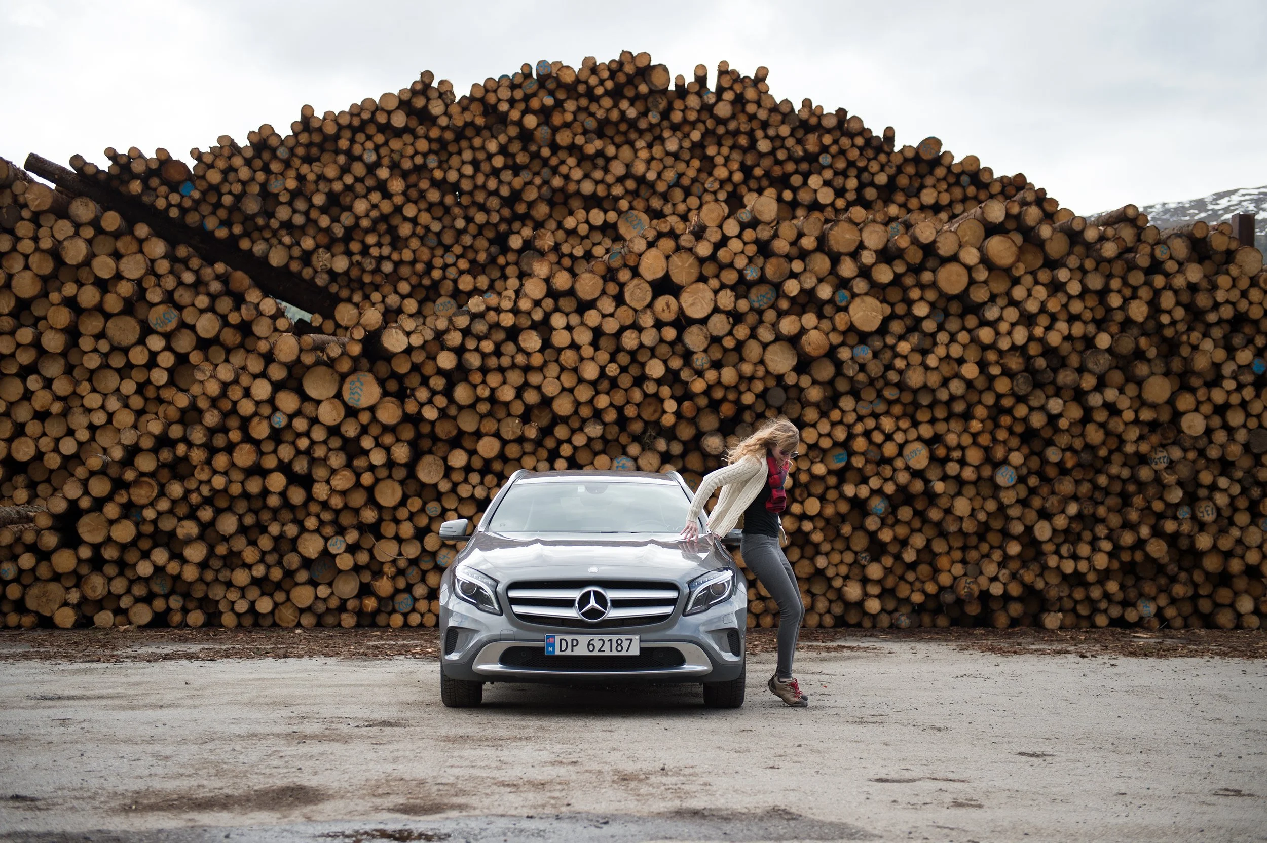 a woman hops onto the hood of a mercedes in front of a log pile in norway