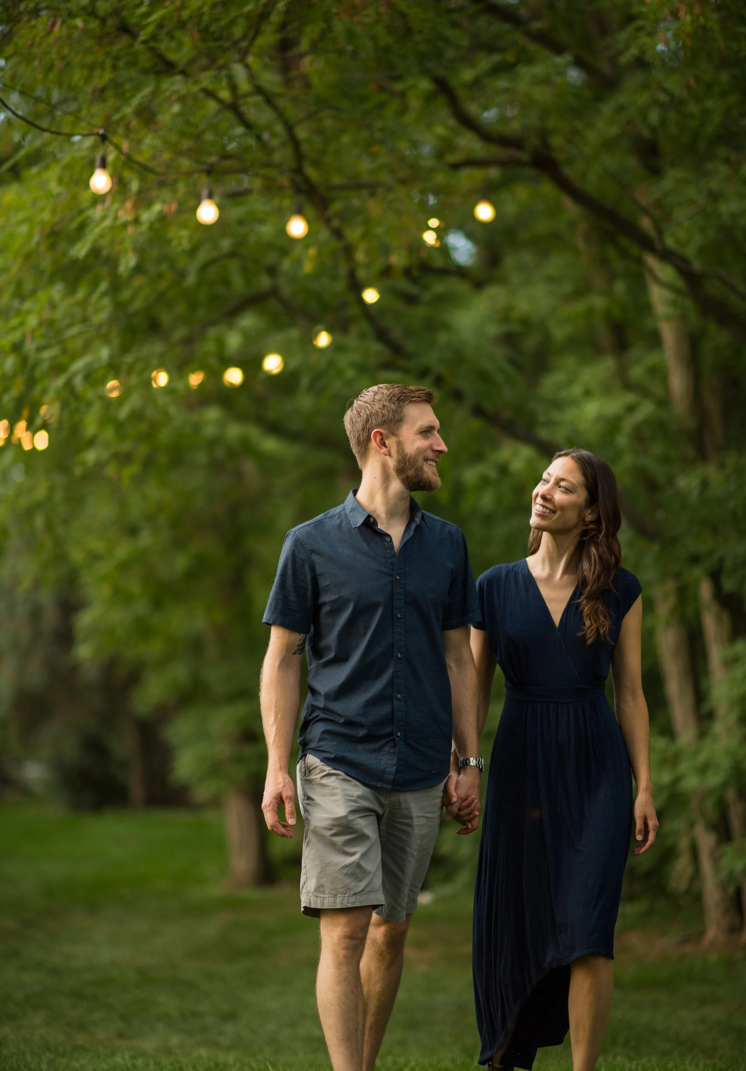 lifestyle photography of boyfriend and girlfriend holding hands while they walk under trees with string lights hanging from them