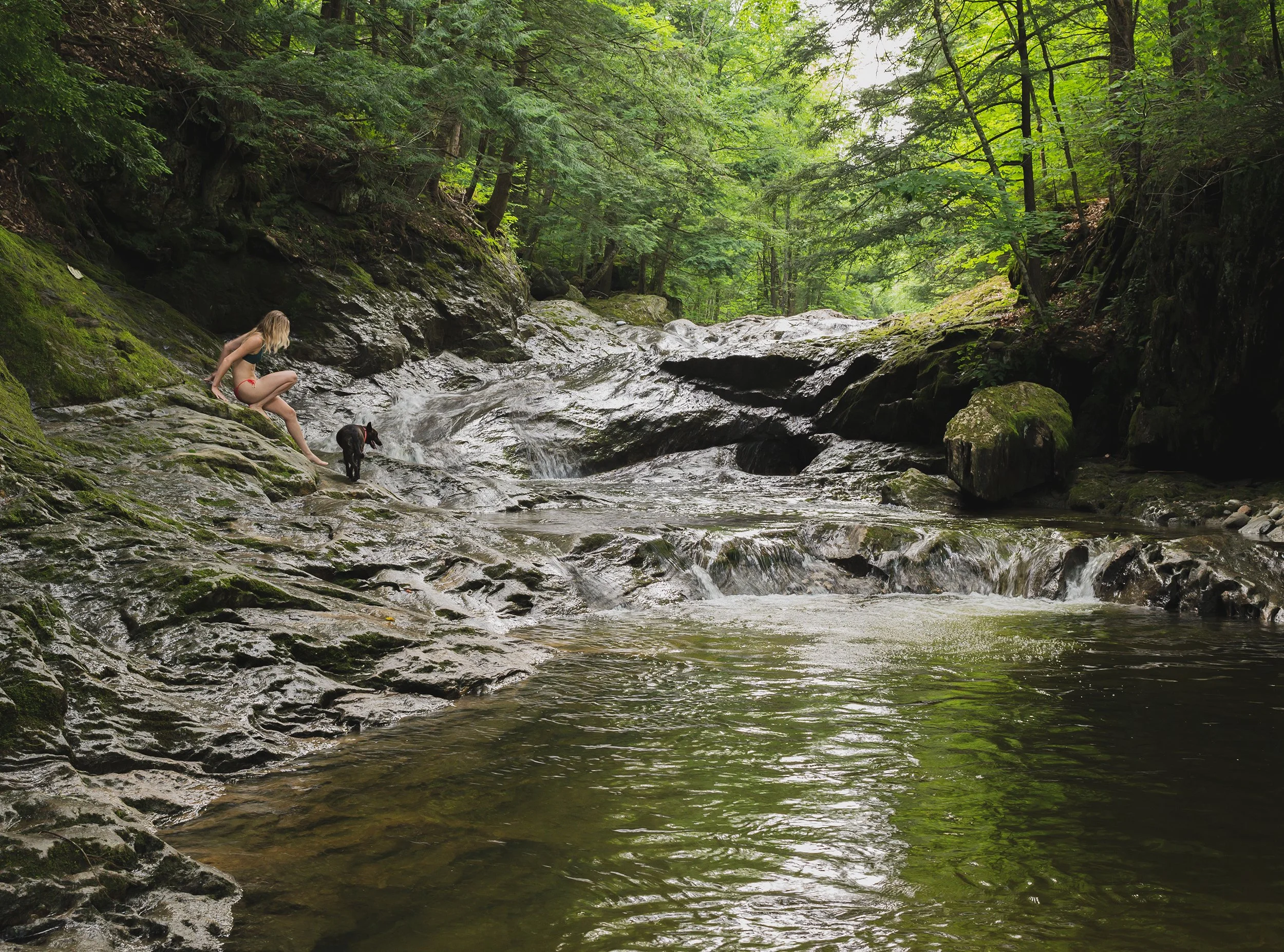 a woman and her dog descend down slippery rocks to a hidden vermont swimming hole