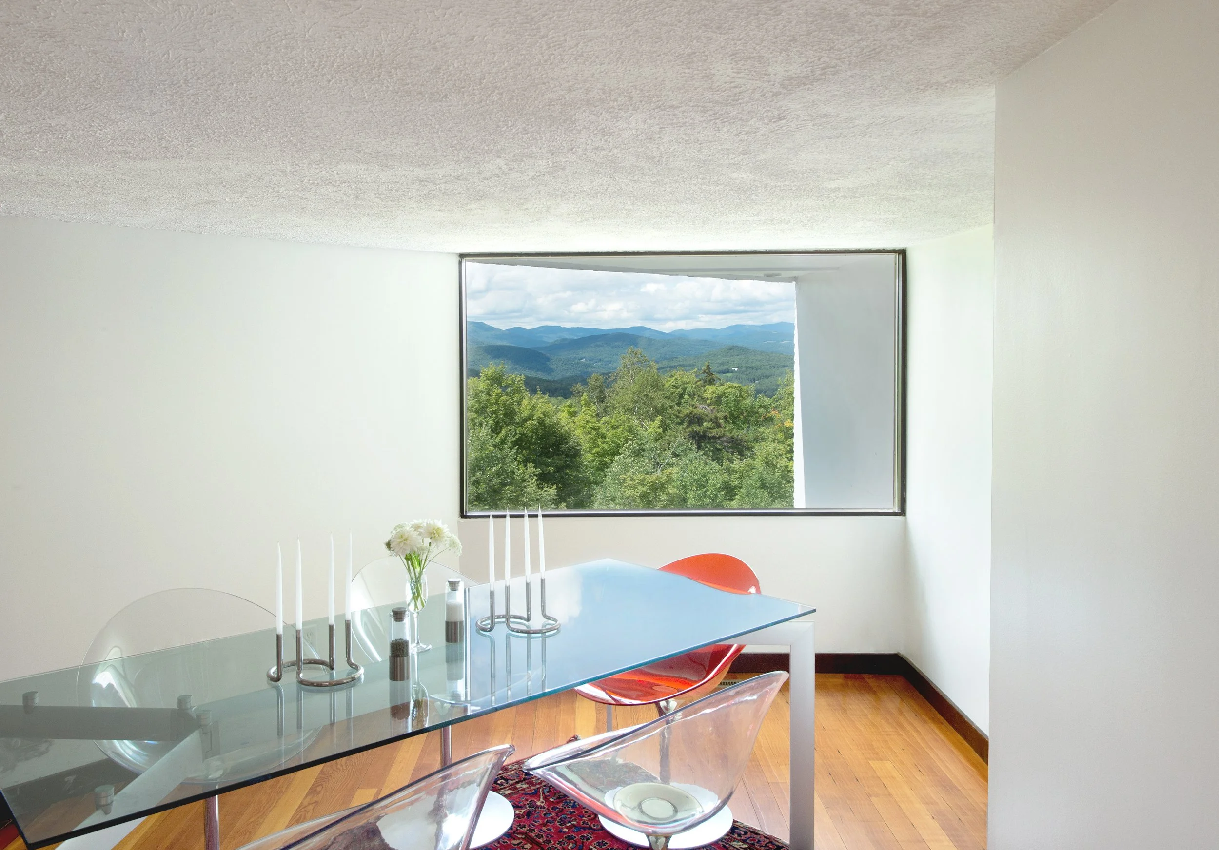 Dining room with glass table, clear and orange chairs, and large window showing a forest and mountain view.