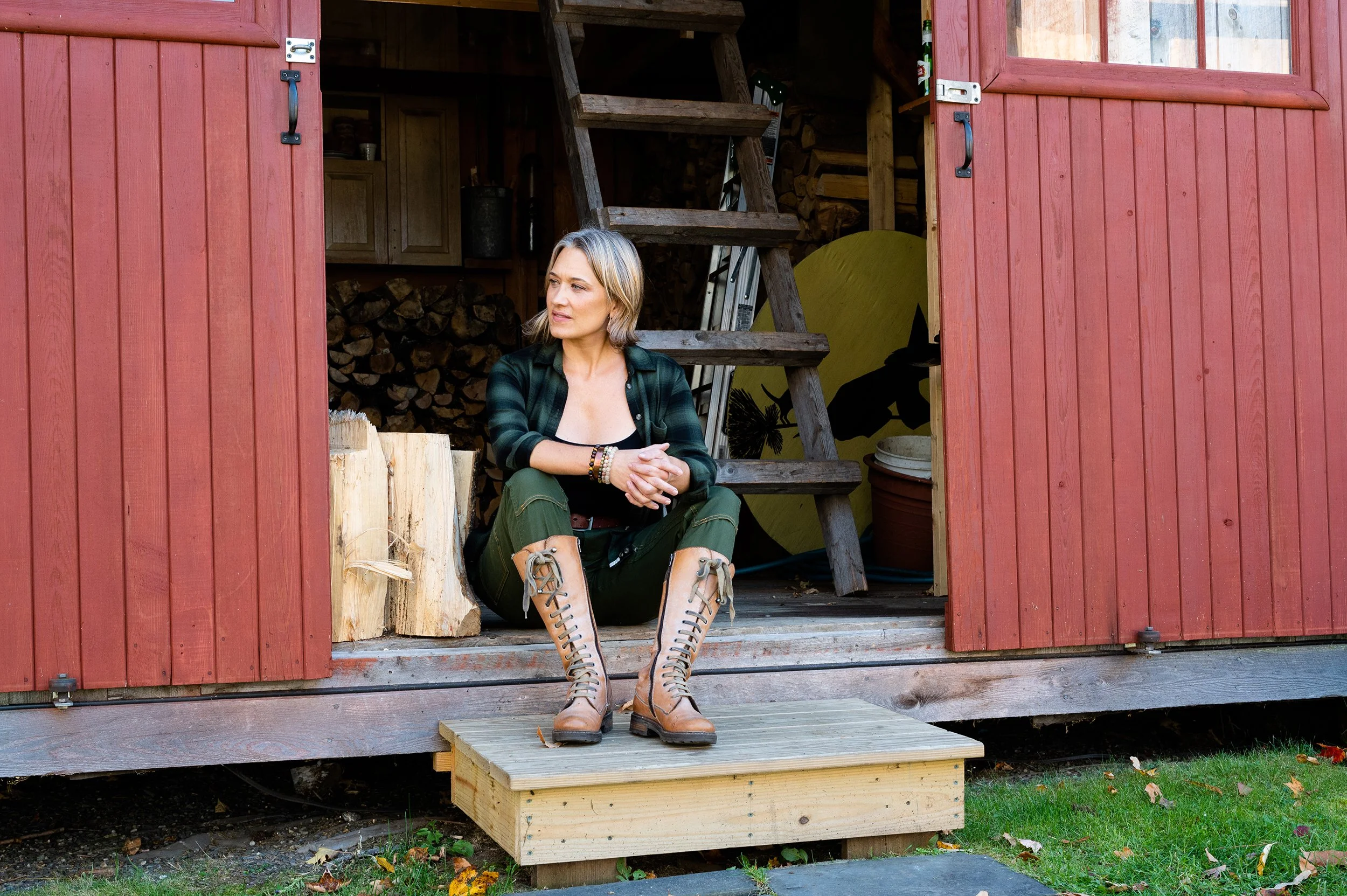 lifestyle photo of woman wearing a green flannel and tall boots sitting on the step of her red barn