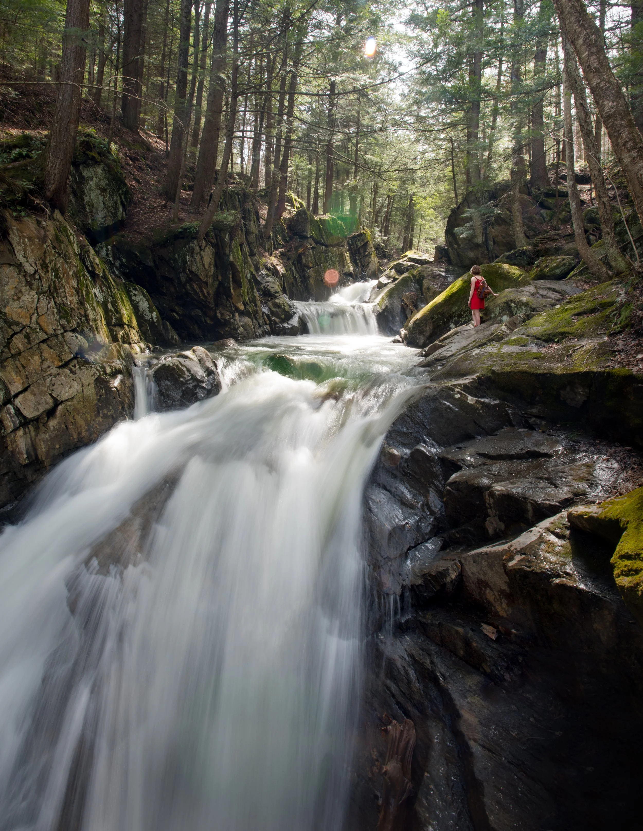 woman in a red dress stands at the top of a waterfall in a vermont forest