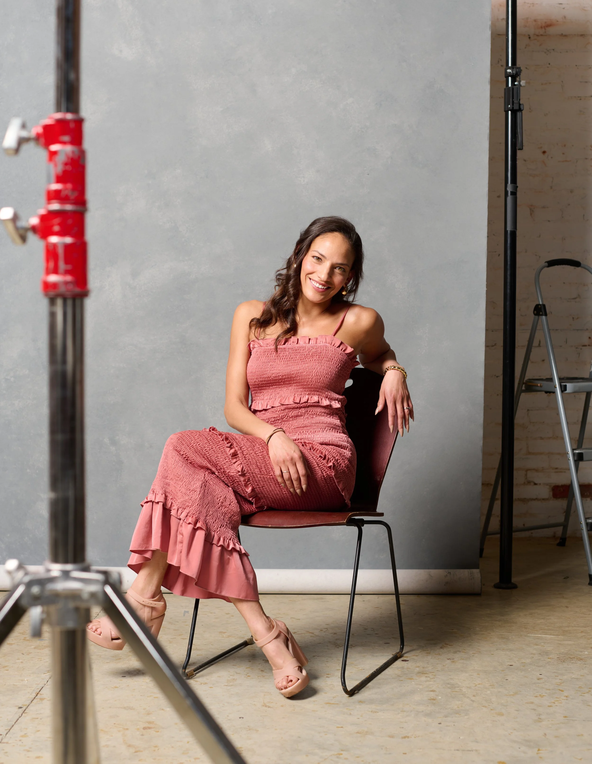behind the scenes photo of melissa bates of meliority house in a red dress sitting on a wooden chair in a photography studio during a vermont fashion photo shoot