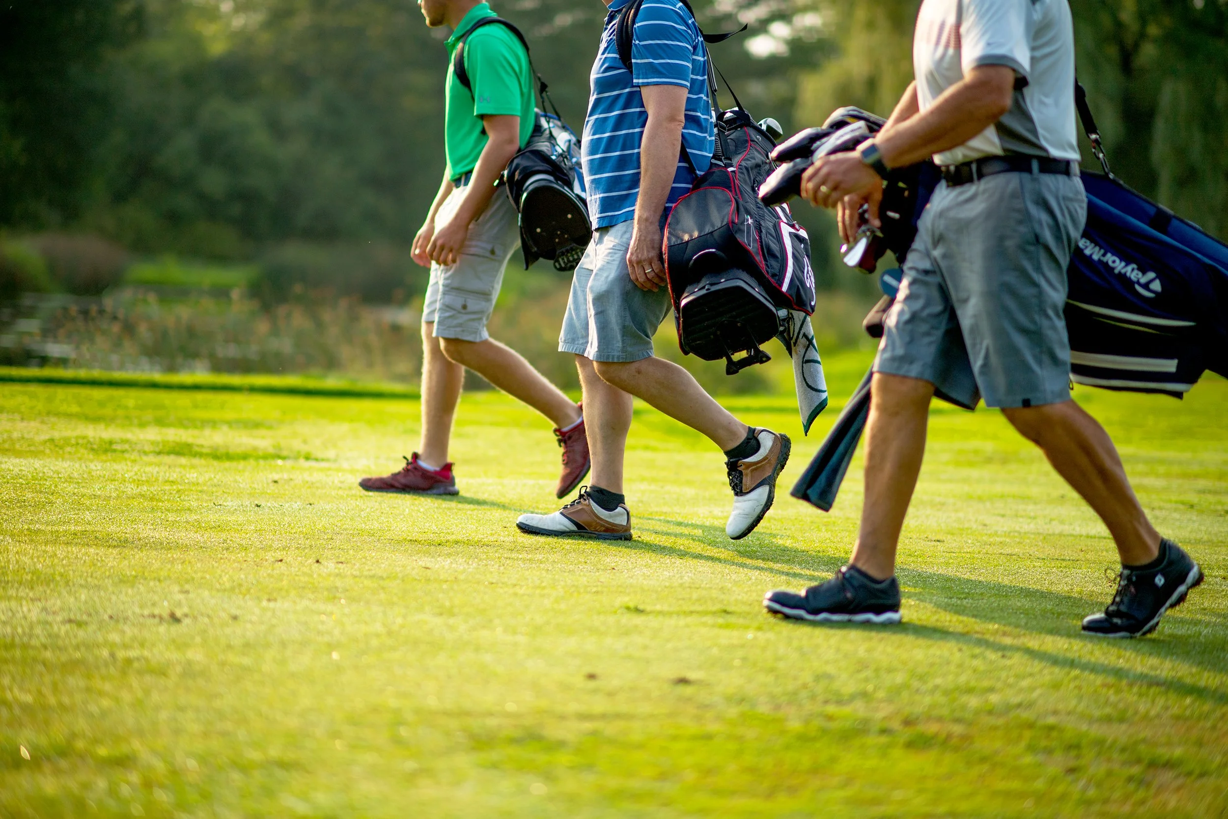 lifestyle photography of three men wearing golf attire and carrying golf club bags walk down a green fairway at basin harbor vermont