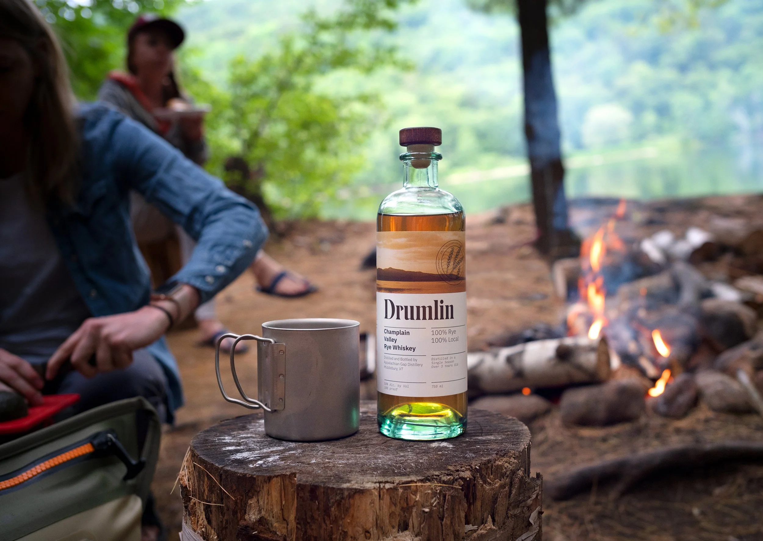 bottle of App Gap Distillery Drumlin Whiskey sitting on a log at a campsite with a campfire in the background