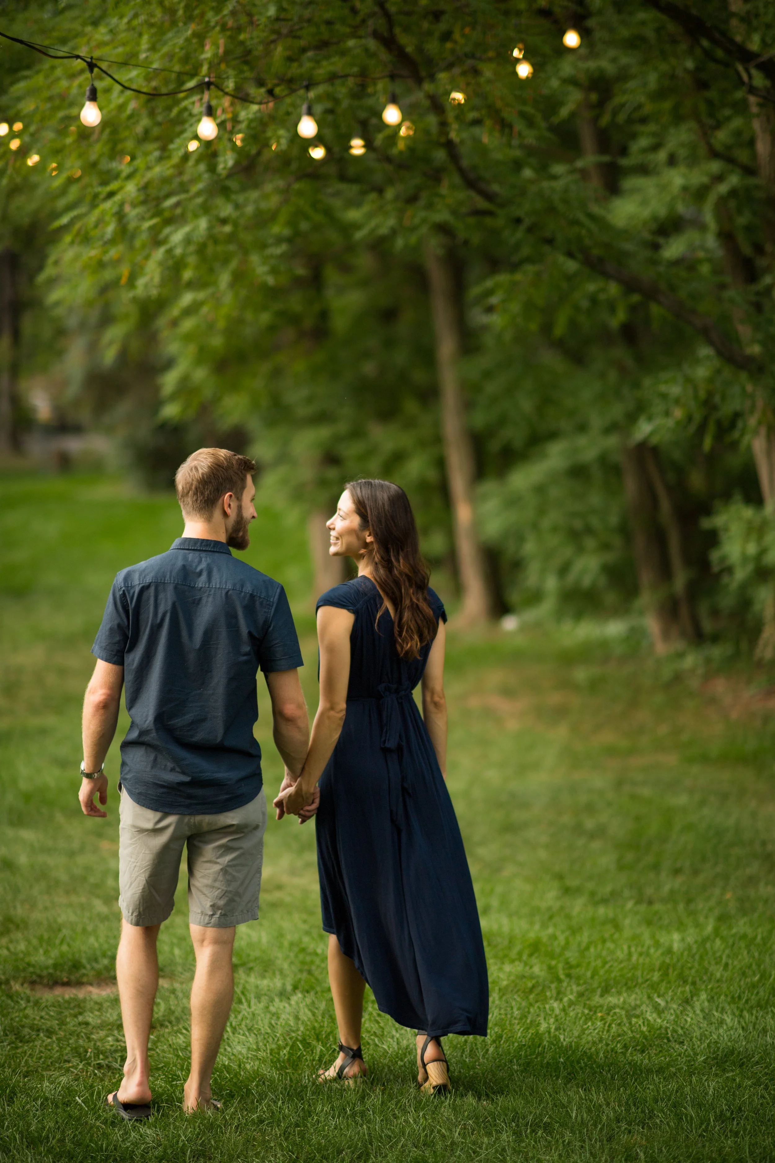 lifestyle photography of man and woman hold ing hands while walking through green grass under green trees with string lights hanging from them