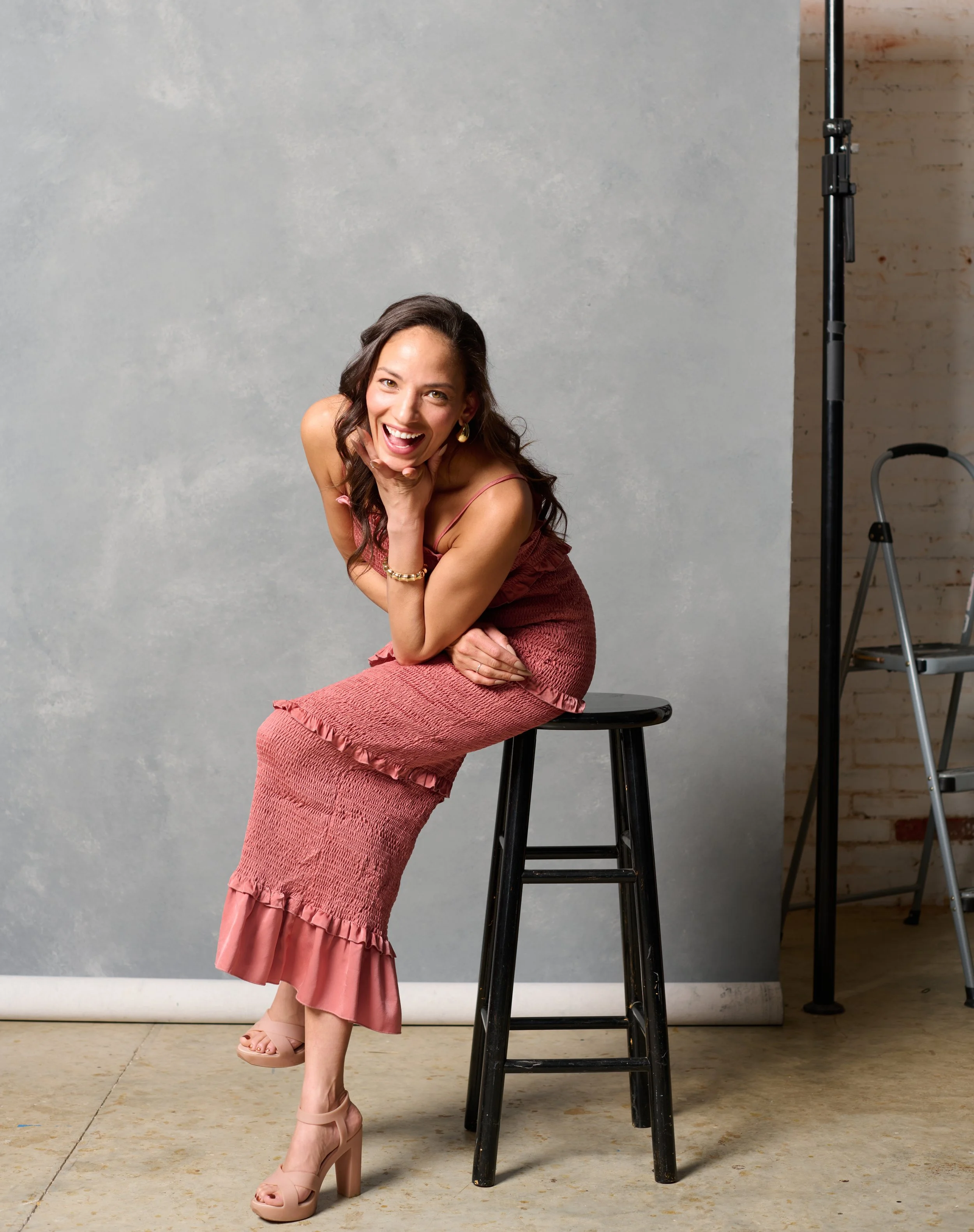 melissa bates of meliority house in a red summery dress laughs as she sits on a stool during a fashion shoot in a Burlington vermont studio