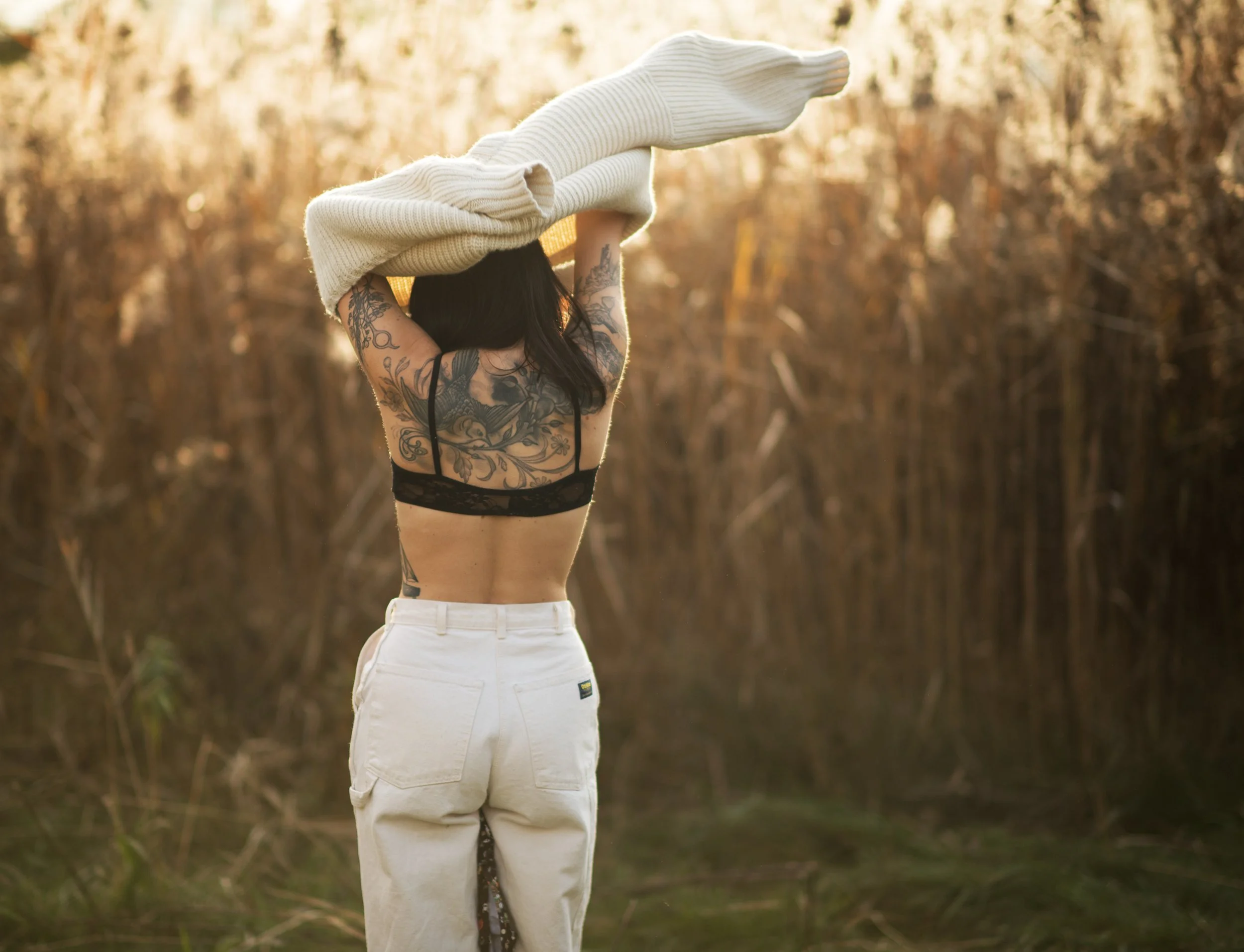 Woman outdoors putting on a sweater, back view, with tattoos on her back.