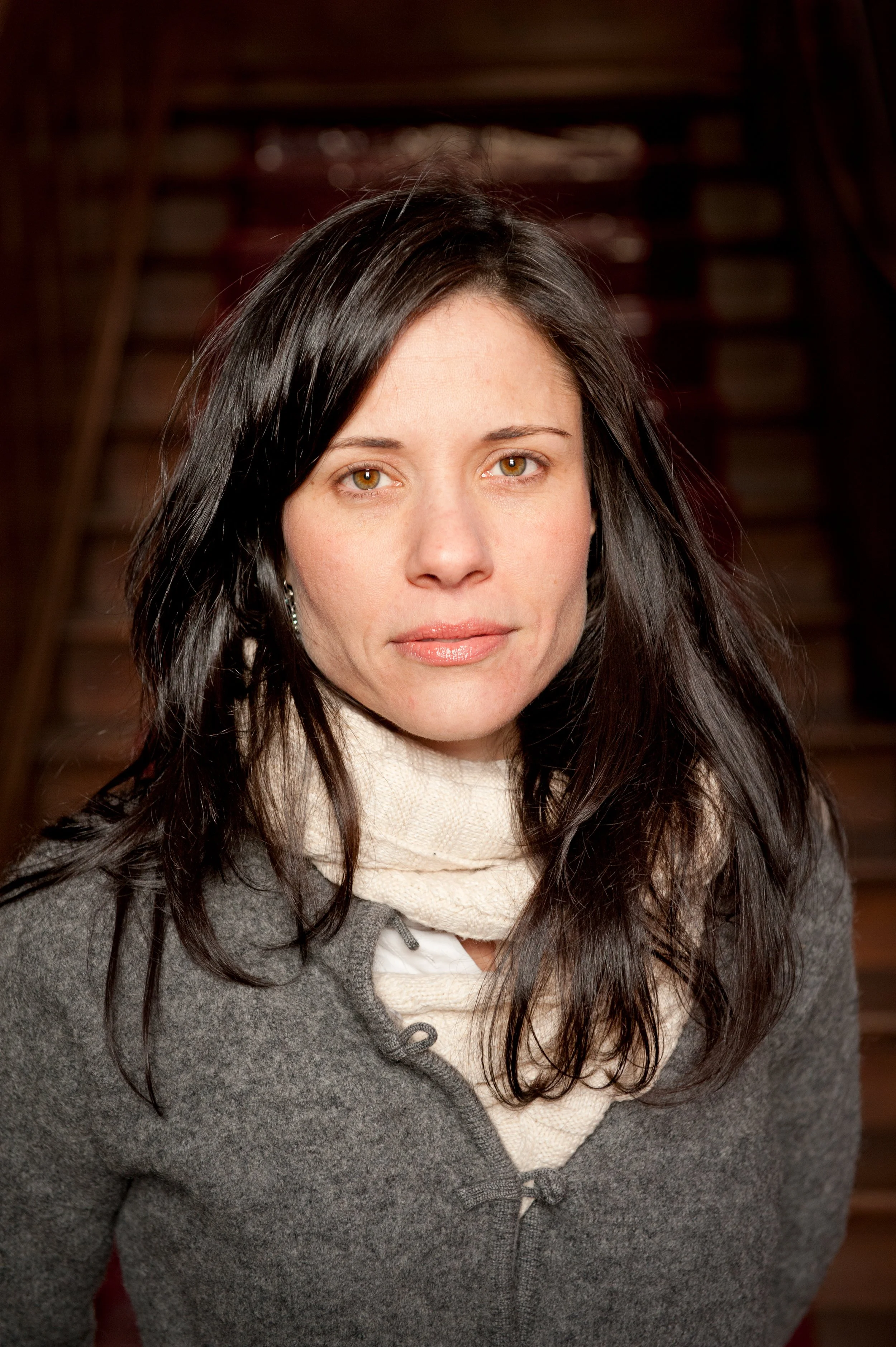 Shannon McNally with black hair wearing a gray sweater and beige scarf, standing indoors with a neutral expression.