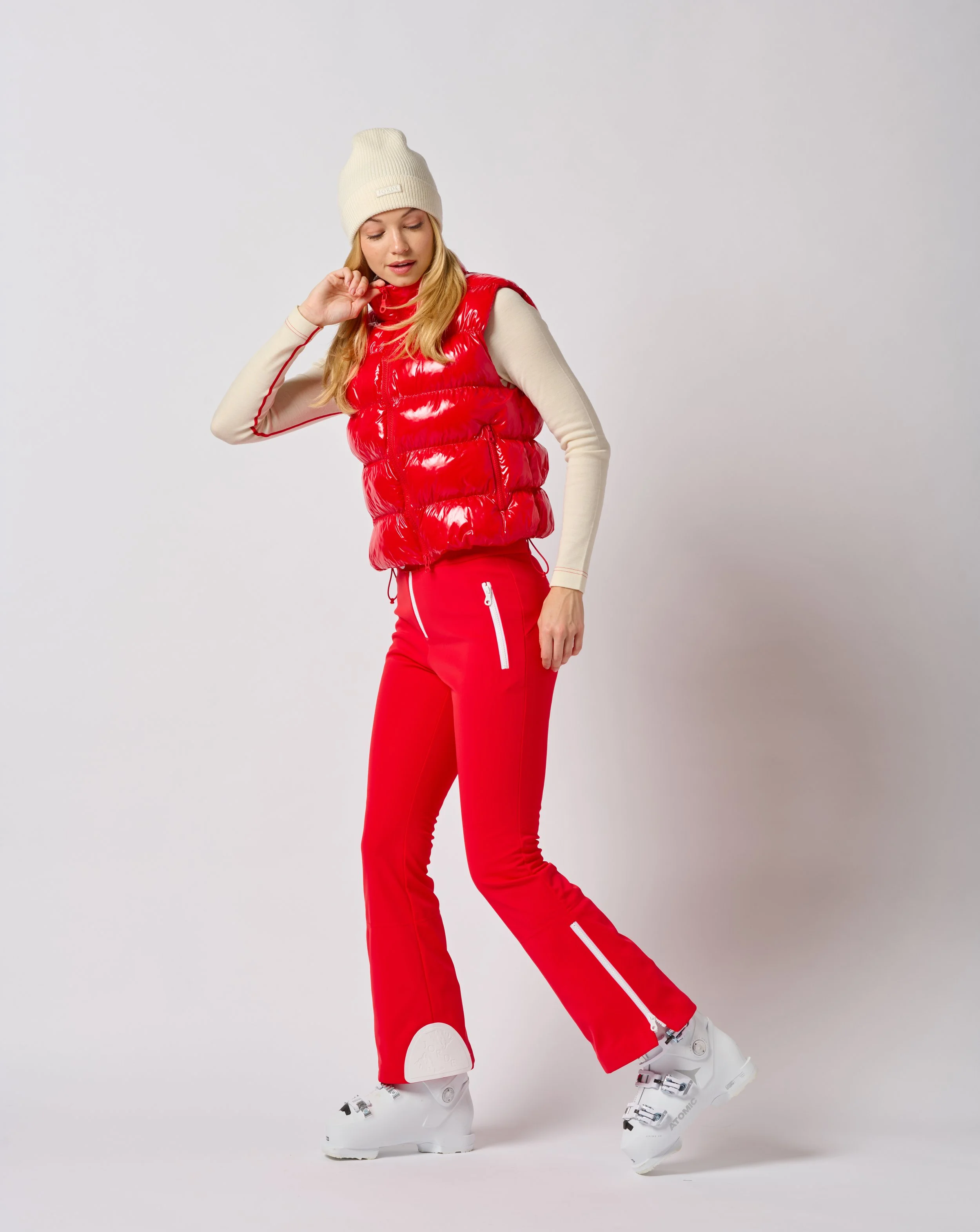 A woman in Jorde Skiwear winter sports attire, wearing a beige beanie, a red puffy vest, beige long-sleeve shirt, red ski pants with white zippers, and white ski boots, standing against a plain white background in a Burlington Vermont photography stu