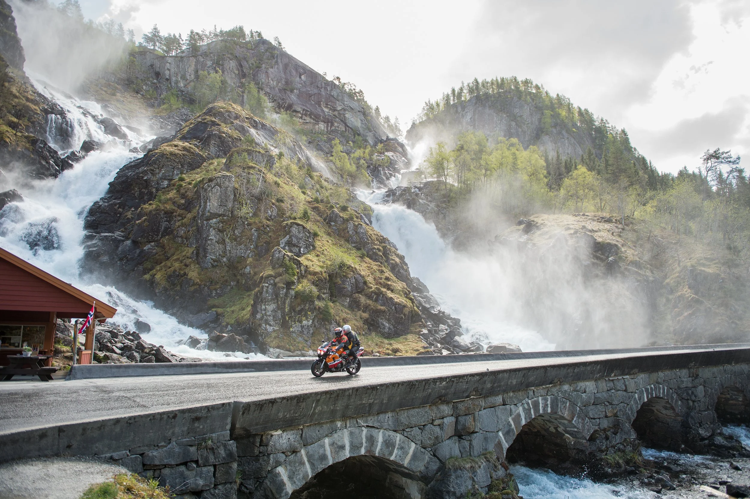 rivers of spring snowmelt flow down cliffs next to a road while two people on a motorcycle ride by