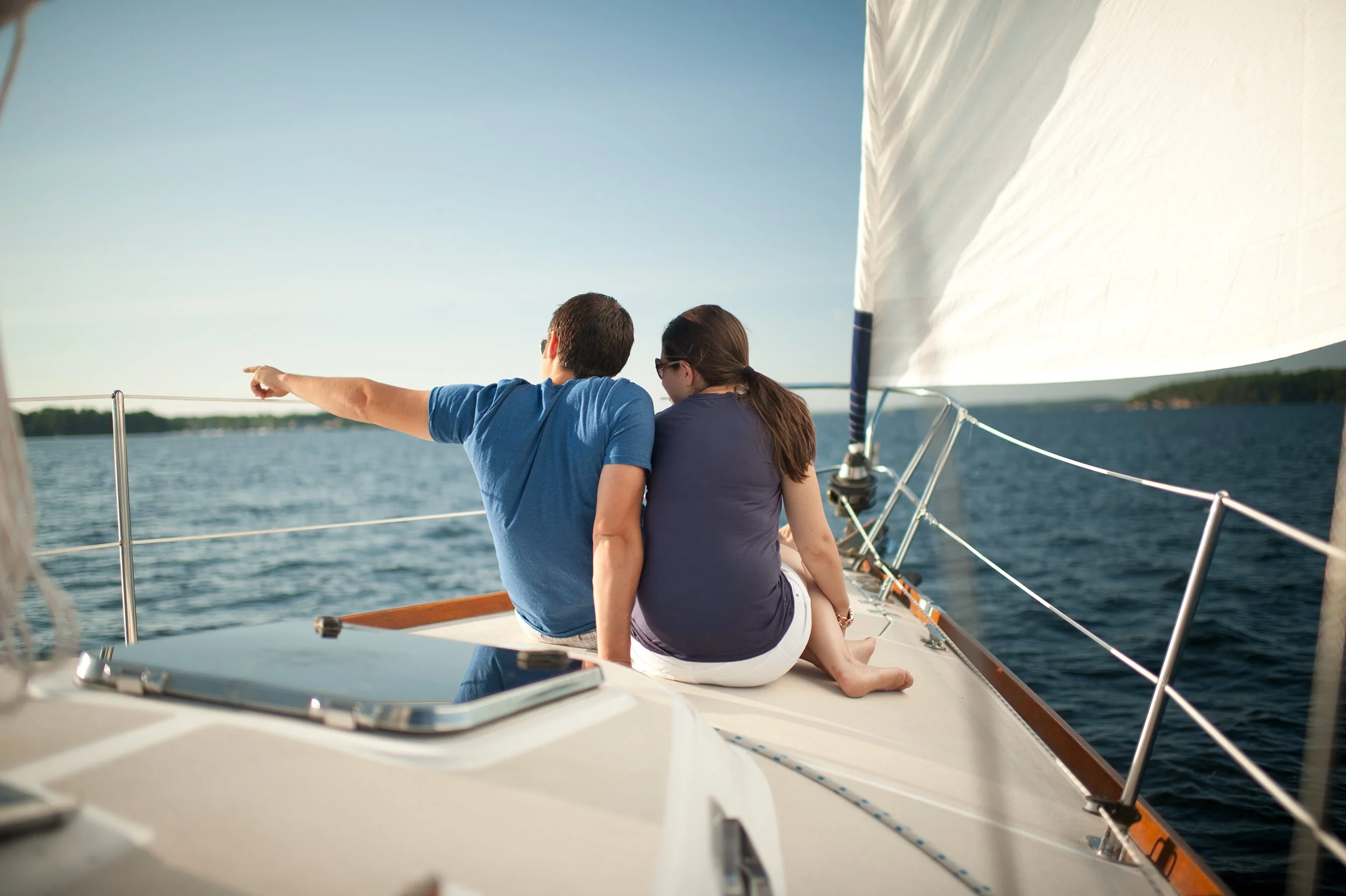 man and woman both wearing blue shirts sit on the bow of a sailboat together as the man points at something in the distance
