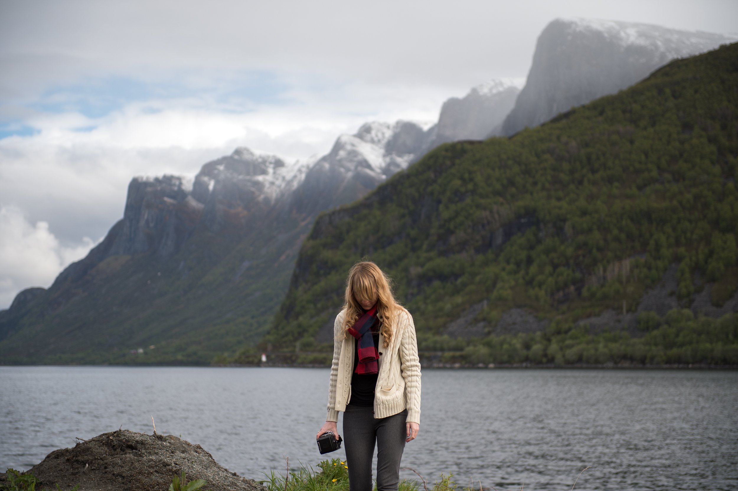a woman in a white sweater and red scarf holds a camera in front of a norwegian fjord and mountains
