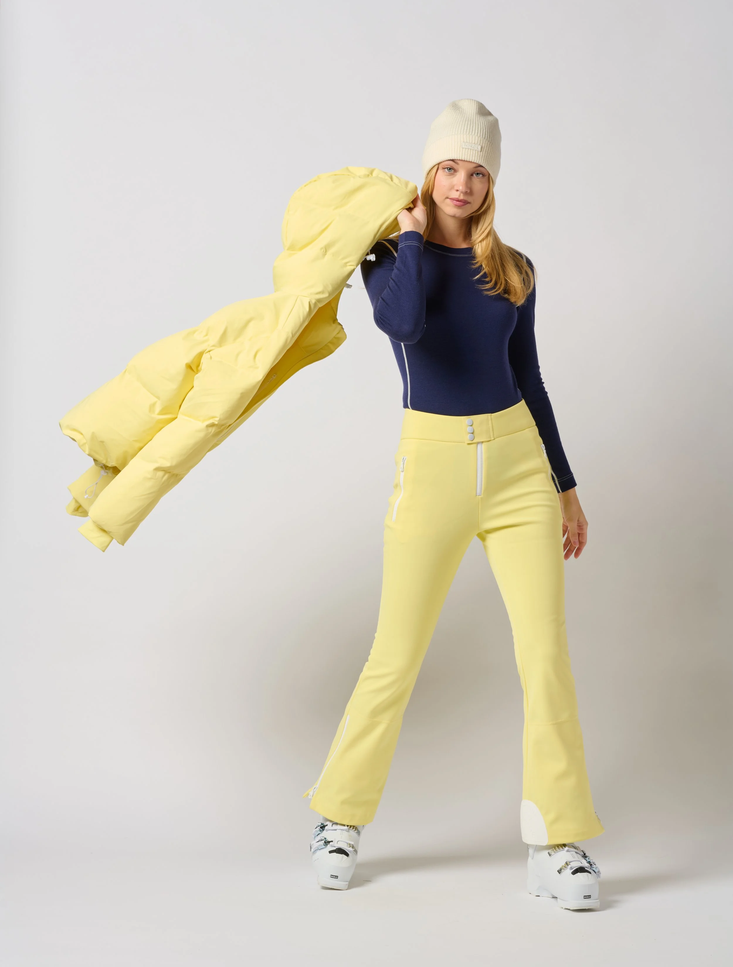 Young woman in yellow Jorde Skiwear ski pants, navy sweater, beanie, and ski boots holding a yellow puffer jacket over her shoulder in a Burlington Vermont photography studio.