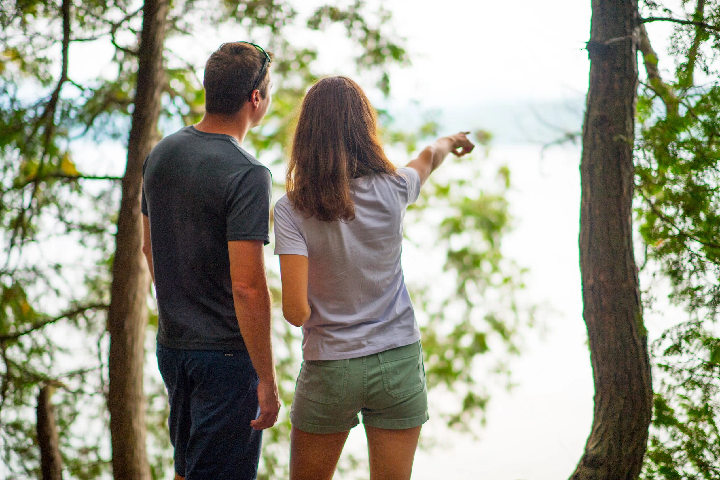 burlington vermont lifestyle photographer image of a female hiker pointing through the trees at lake champlain while her boyfriend looks at where she is pointing