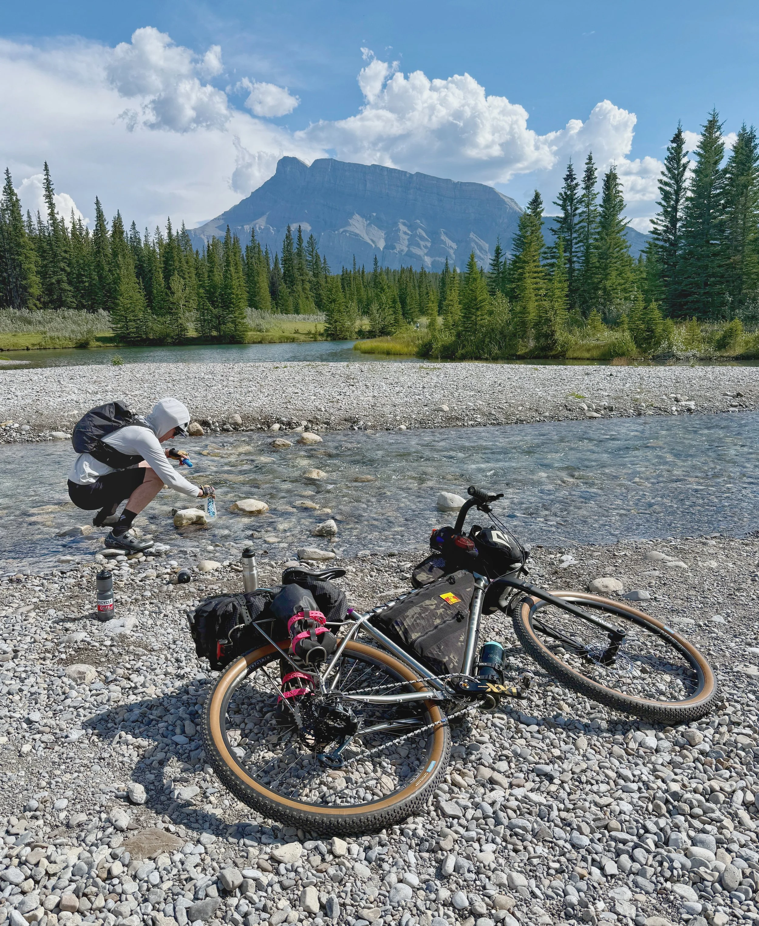 A person in outdoor gear filling water bottles at a riverbank in Banff national park with a mountain, trees, and a partly cloudy sky in the background. A bikepacking bicycle is on the rocky shore.