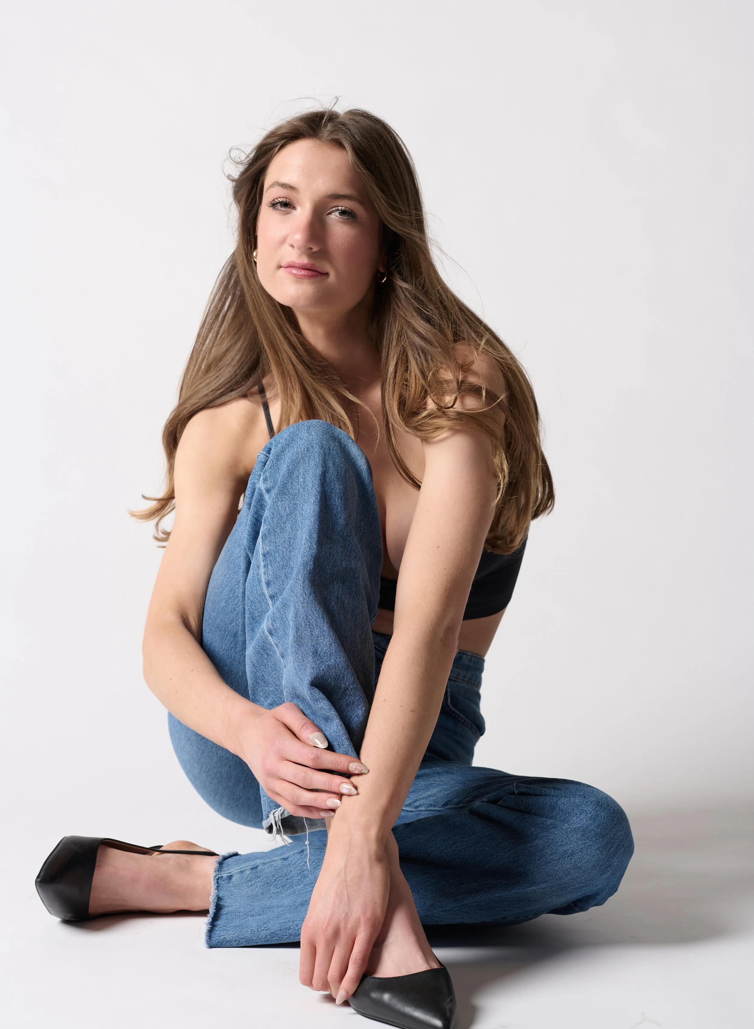 Female model posing for a studio photoshoot on the floor wearing casual denim jeans and black top, with long hair and black heels.