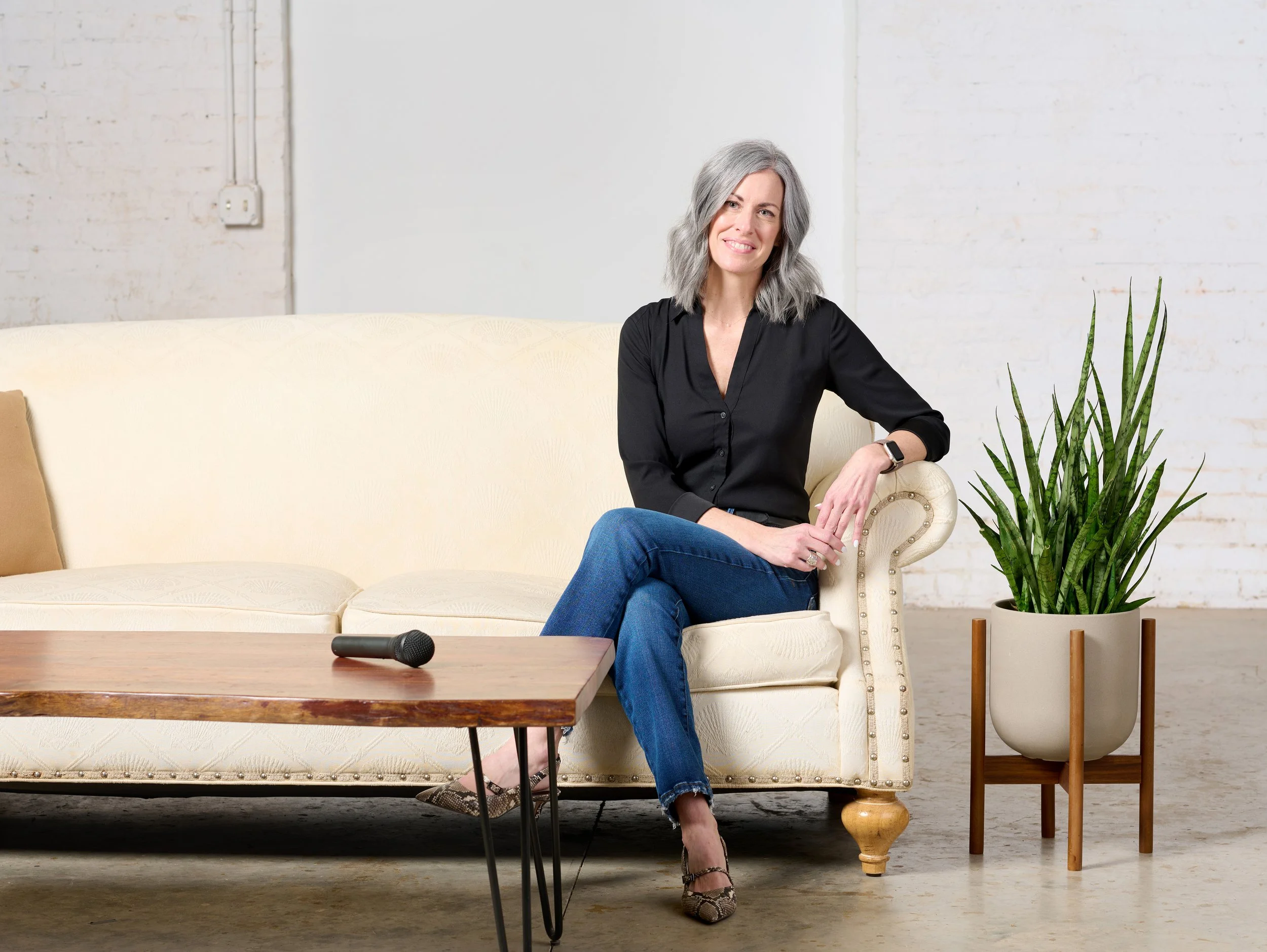 Meg McKeen sits for a studio lifestyle portrait on a cream colored couch next to a green plant with a microphone on table in front of her