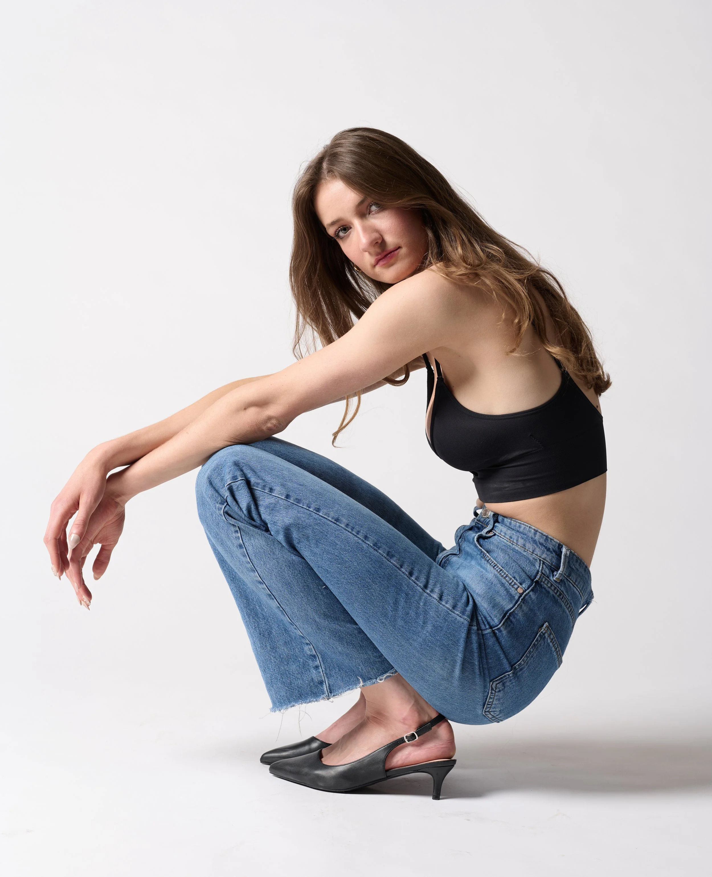 Female model posing for a studio photoshoot while squatting wearing a black crop top, blue jeans, and black heels, with long brown hair, against a white background.