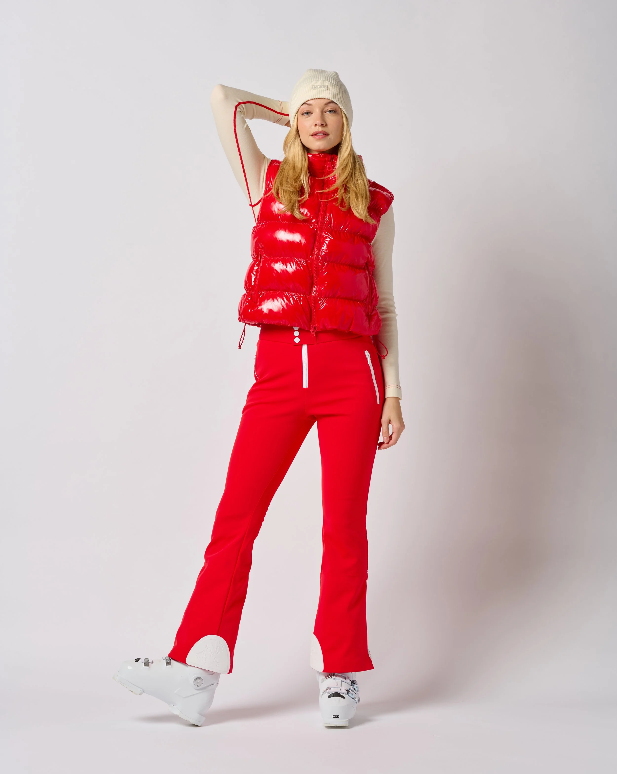 Woman in red ski pants, beige long-sleeve shirt, red puffer vest, white knit hat, and white ski boots posing with one arm behind her head in a in a Burlington Vermont photography studio.