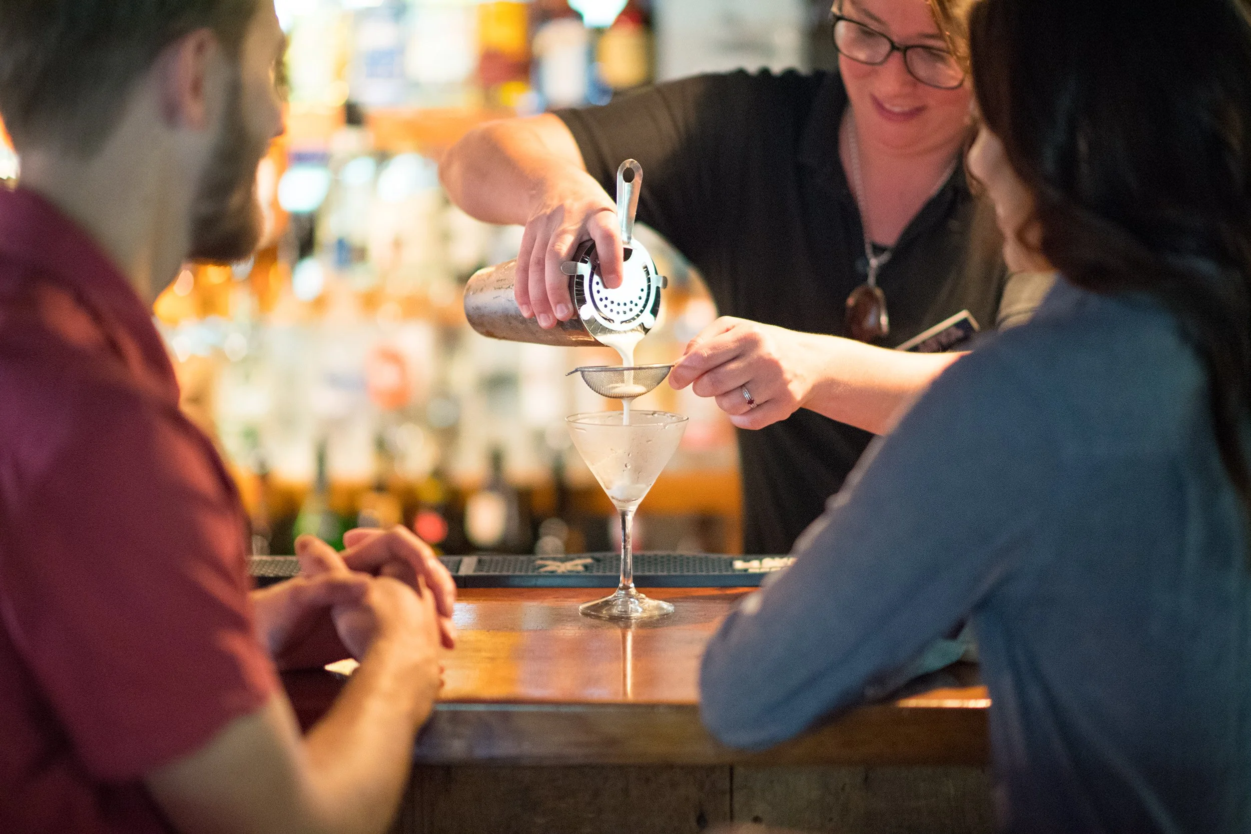 a bartender pours a cocktail in to a martini glass while two patrons look on