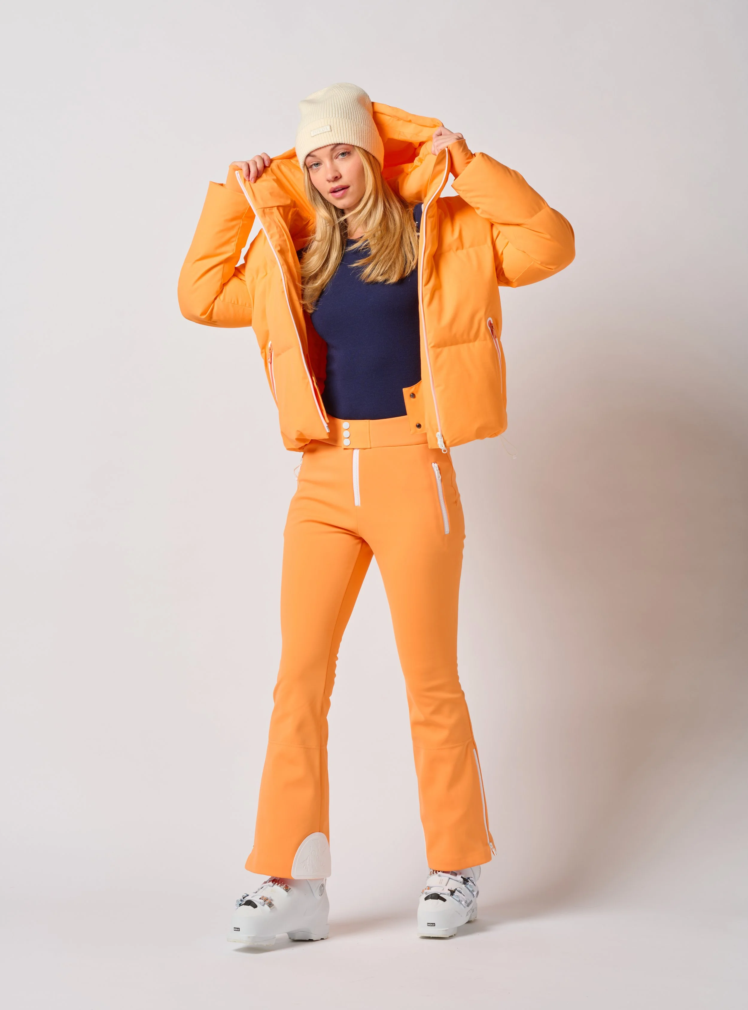 A woman dressed in Jorde Skiwear winter ski gear, wearing an orange puffer jacket, orange pants, a beige beanie, and white ski boots, standing against a plain white background in a Burlington Vermont photography studio.