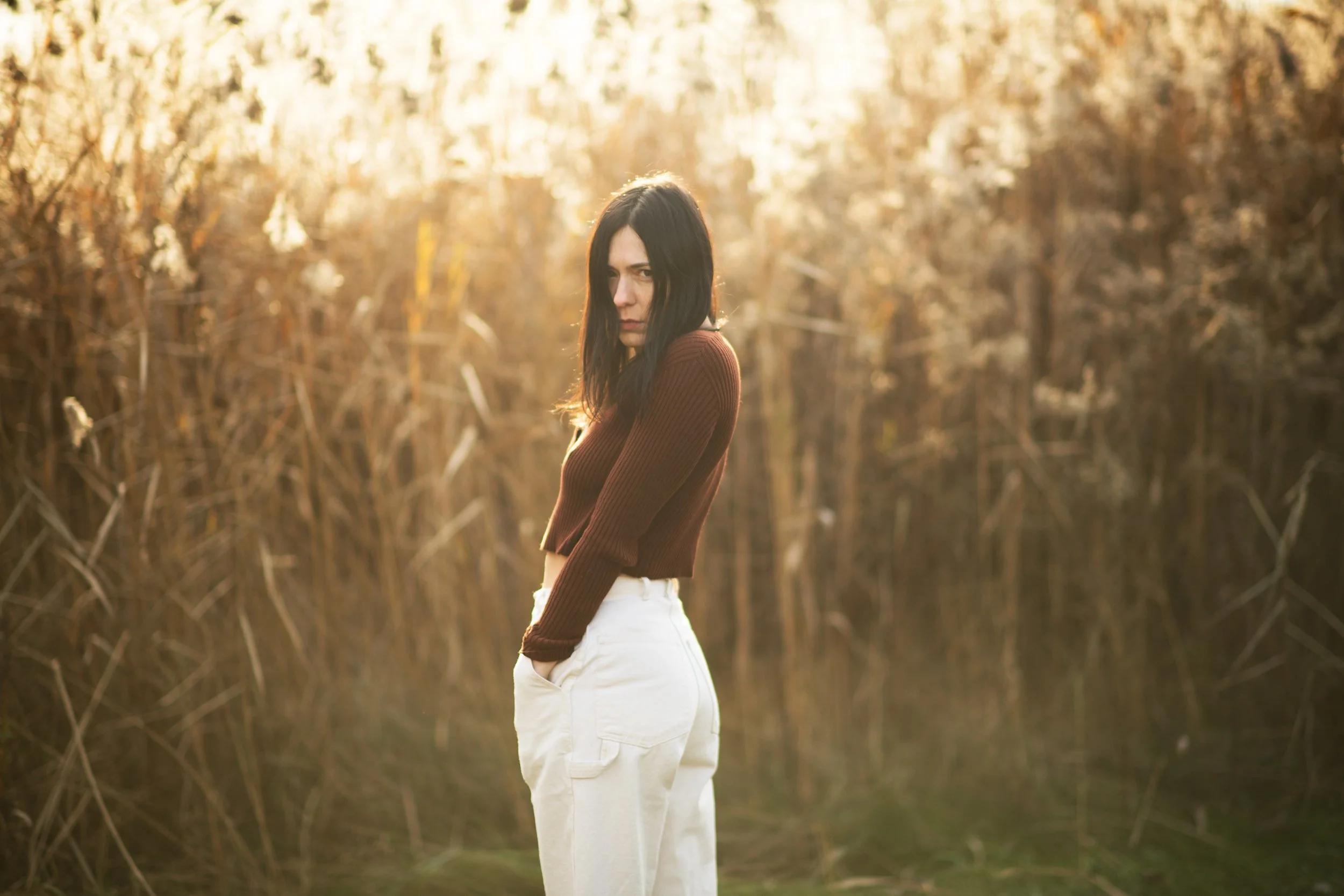Woman standing in tall grass field wearing a brown sweater and white pants, looking pensive.