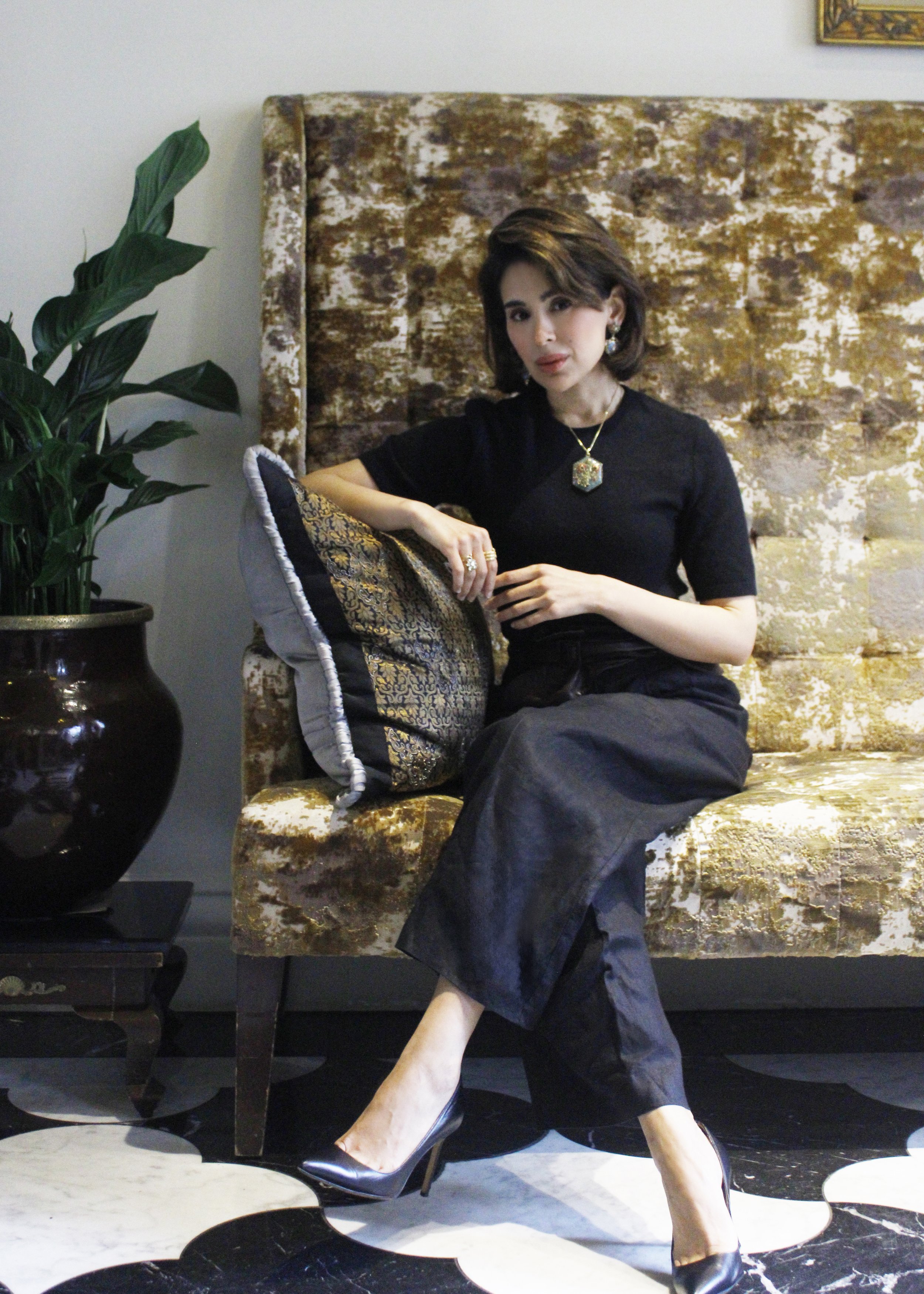 Woman sitting on a gold-patterned couch in front of a textured gold and white wall, wearing a black dress and high heels, with a large green plant nearby.