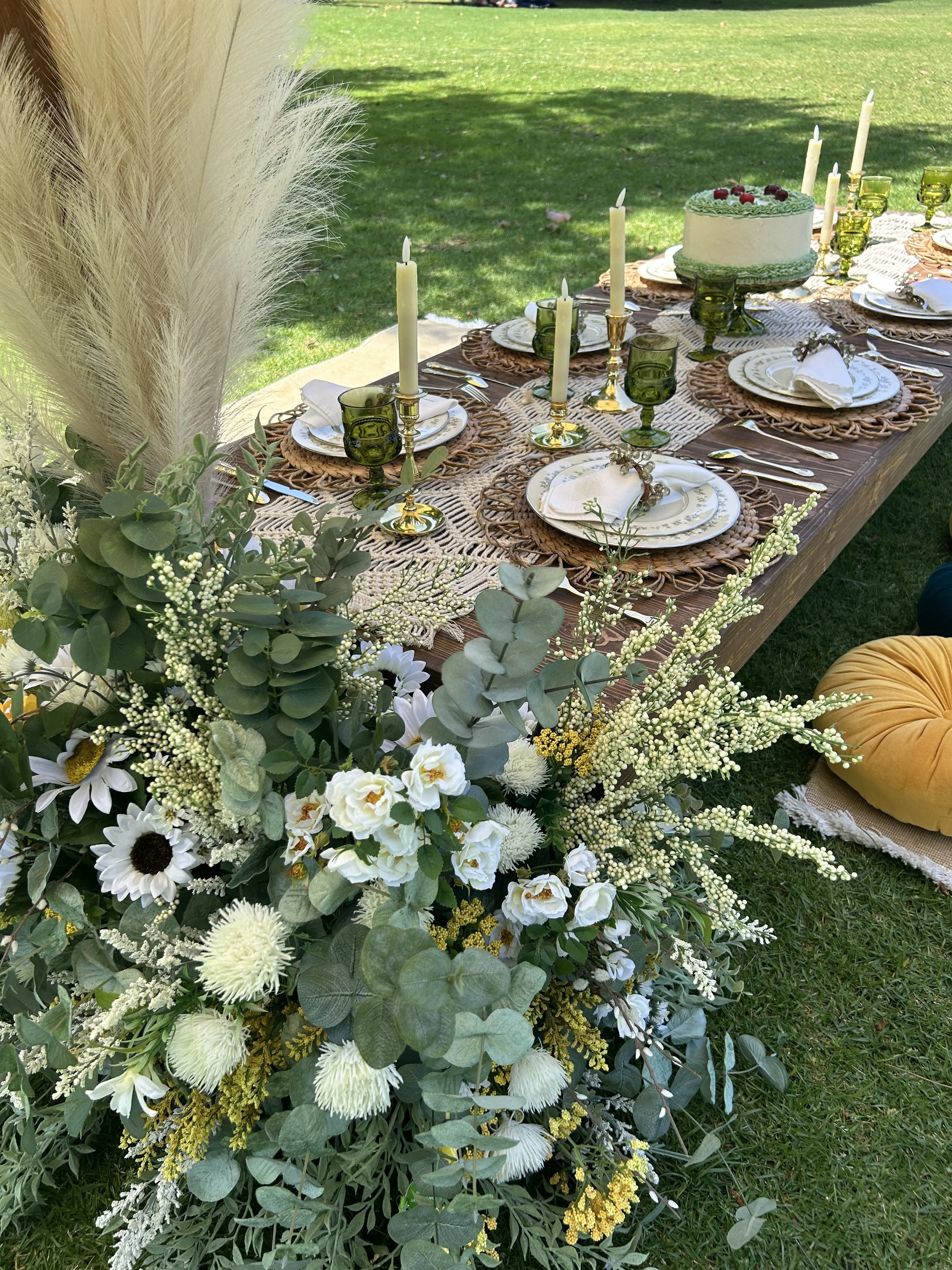 Elegant outdoor picnic table setting with green glassware, gold candle holders, and floral centerpiece featuring white and yellow flowers.