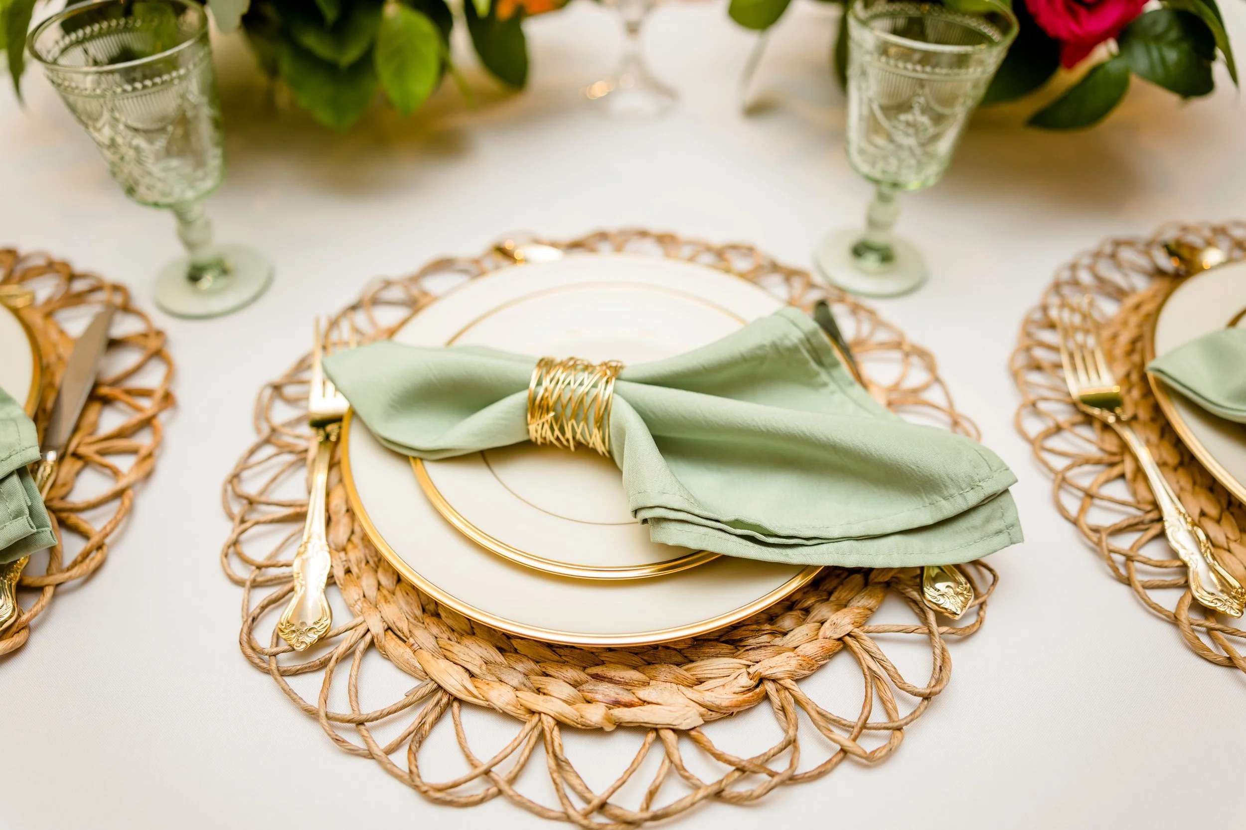 Elegant table setting with a white tablecloth, gold-rimmed plates, and green cloth napkins wrapped with gold rings on woven placemats. Green glassware and floral arrangements are in the background.