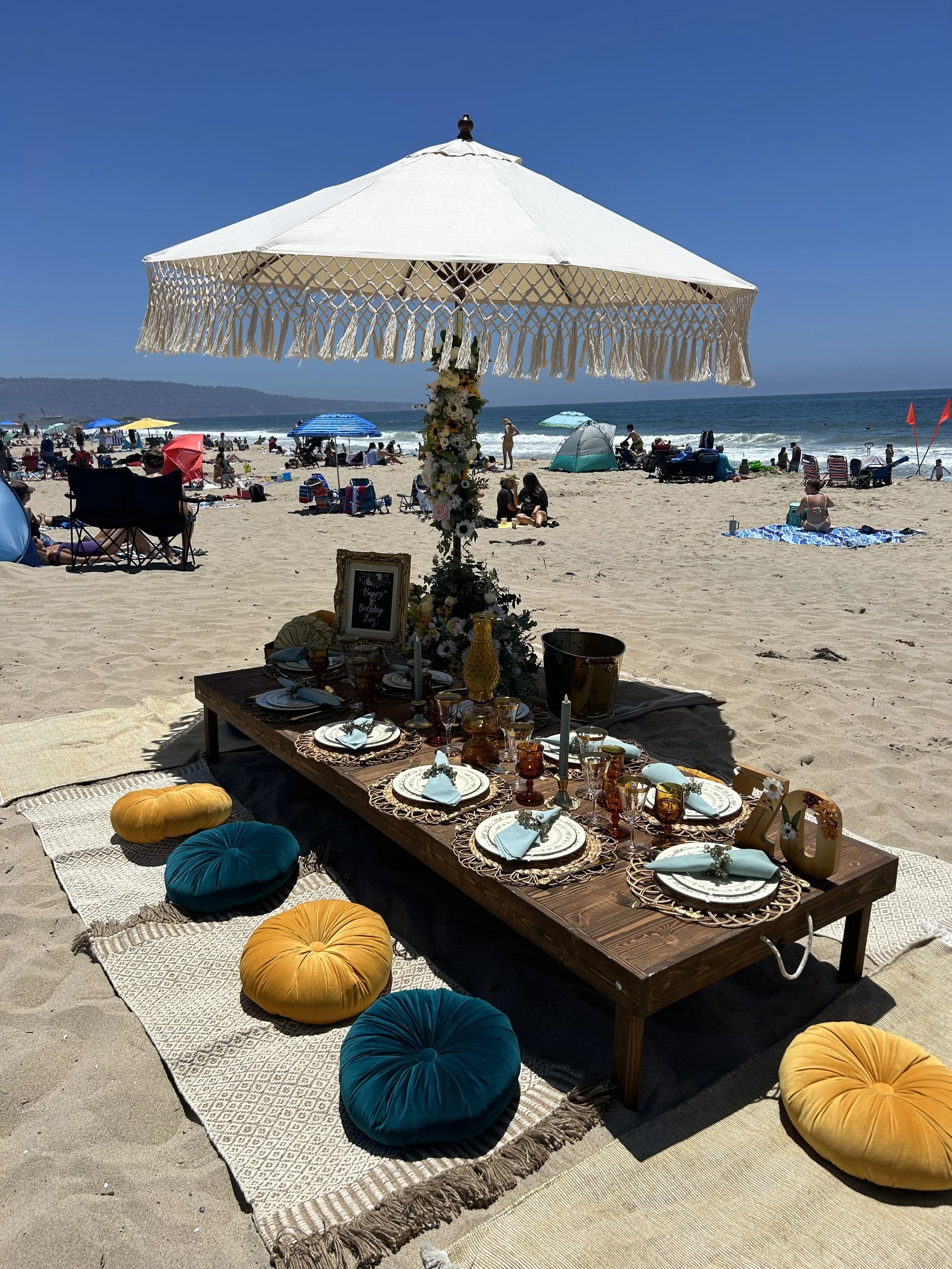 Beach picnic setup with a low wooden table, decorated with plates, glasses, and napkins. The table is surrounded by teal and yellow cushions on a woven mat. A large fringed umbrella provides shade, and people with beach umbrellas can be seen in the background.