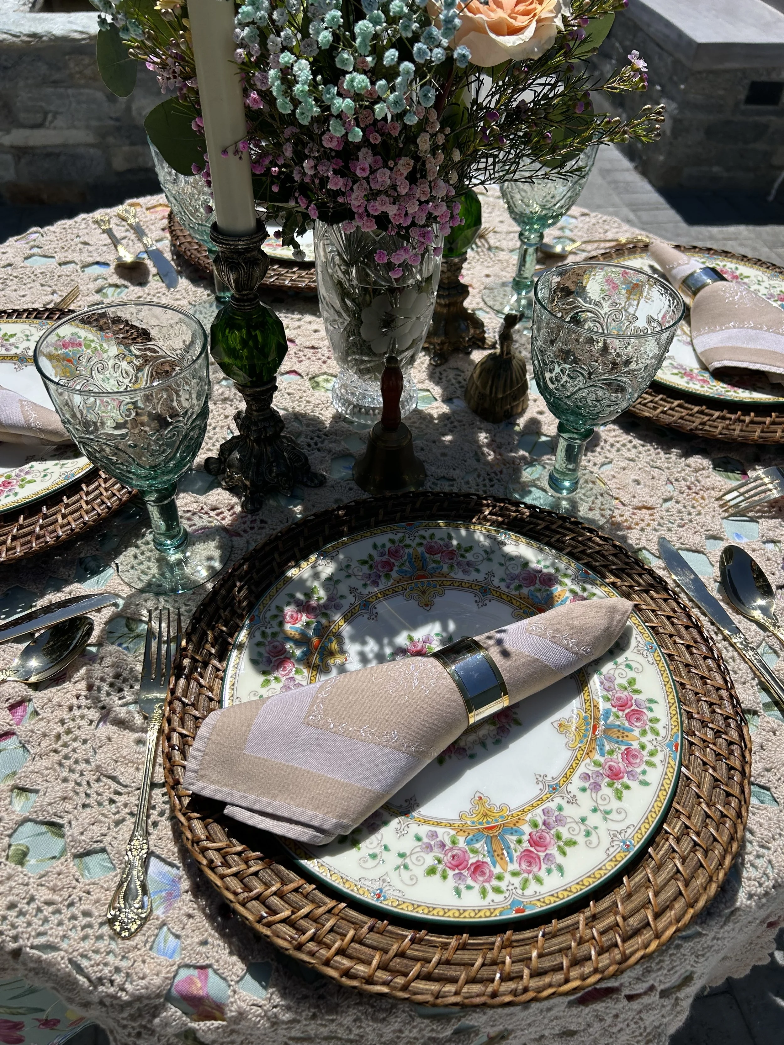 Elegant table setting with floral china plates, wicker charger, embroidered napkins, ornate silverware, crystal glassware, centerpiece of pastel flowers, and lace tablecloth.