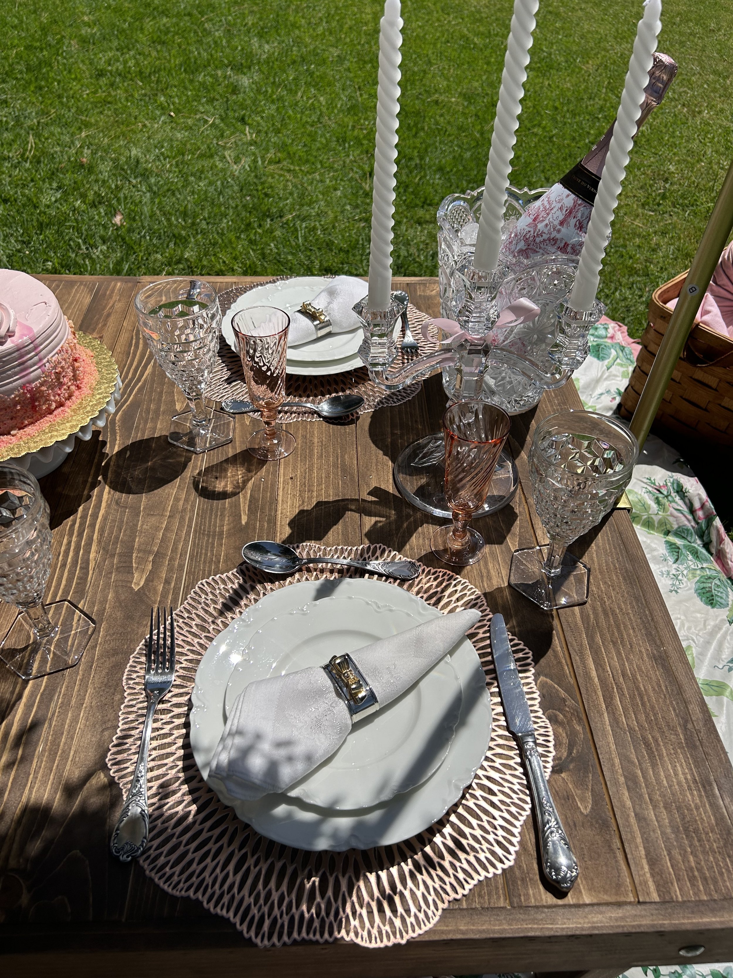 Elegant outdoor table setting with pastel-colored elements, featuring a white plate, intricately folded napkin with a napkin ring, crystal glassware, ornate cutlery, a birthday cake, and tall white candlesticks on a wooden table.