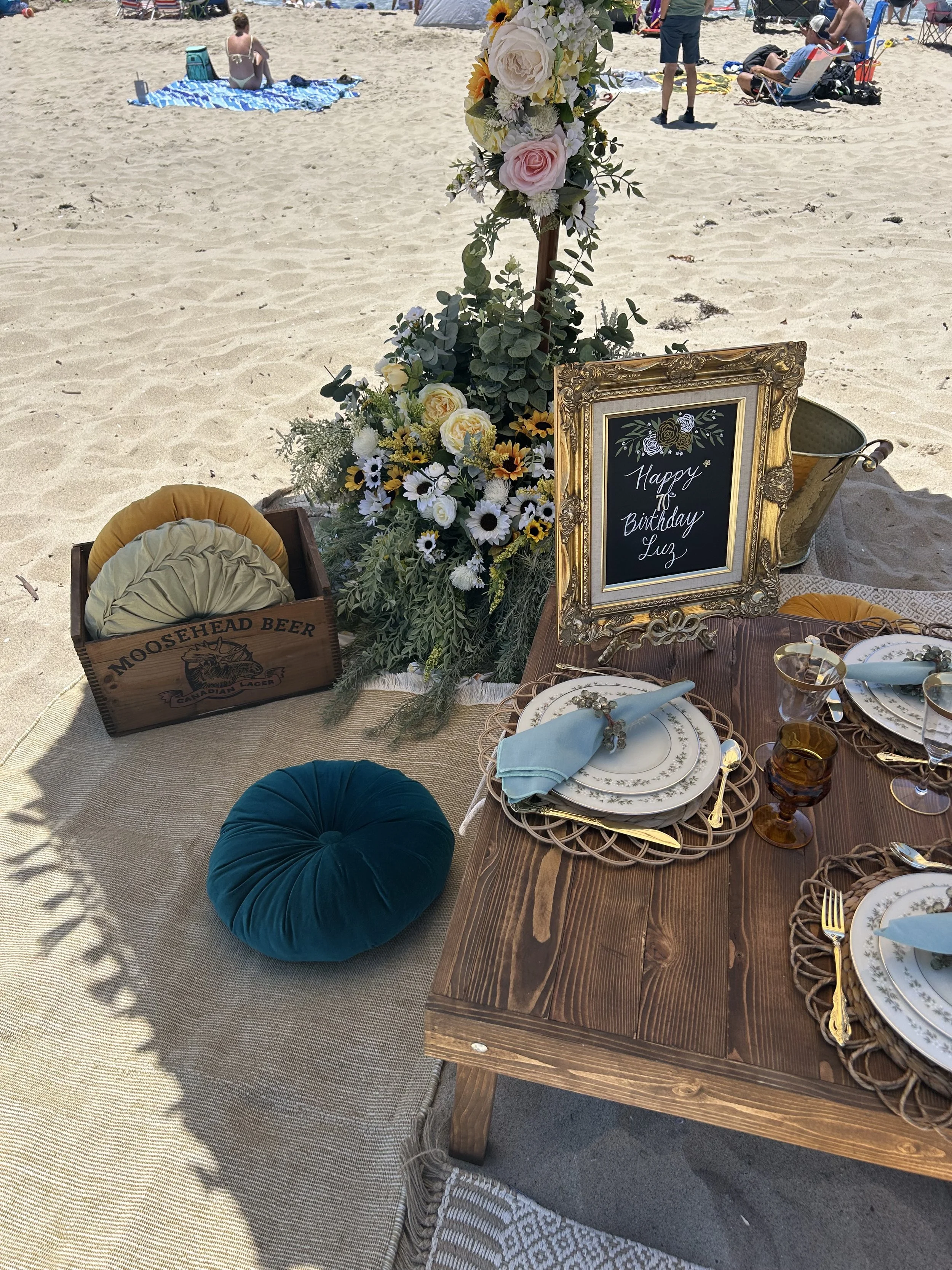 Beach picnic setup with a decorated table, plates, cutlery, and glassware on a wooden table. A bouquet of flowers and greenery is arranged beside a framed sign saying "Happy Birthday Luz." A wooden crate with cushions is nearby, and there are beachgoers in the background.
