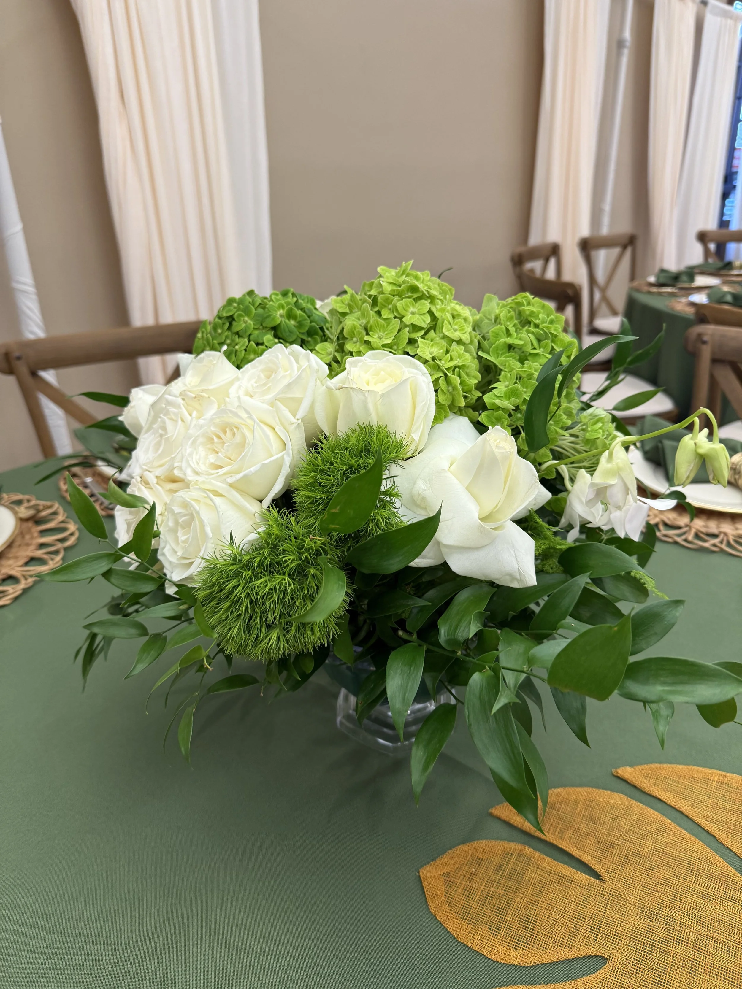 A floral centerpiece with white roses, green hydrangeas, green pompom flowers, and assorted green foliage on a green tablecloth in a dining room.