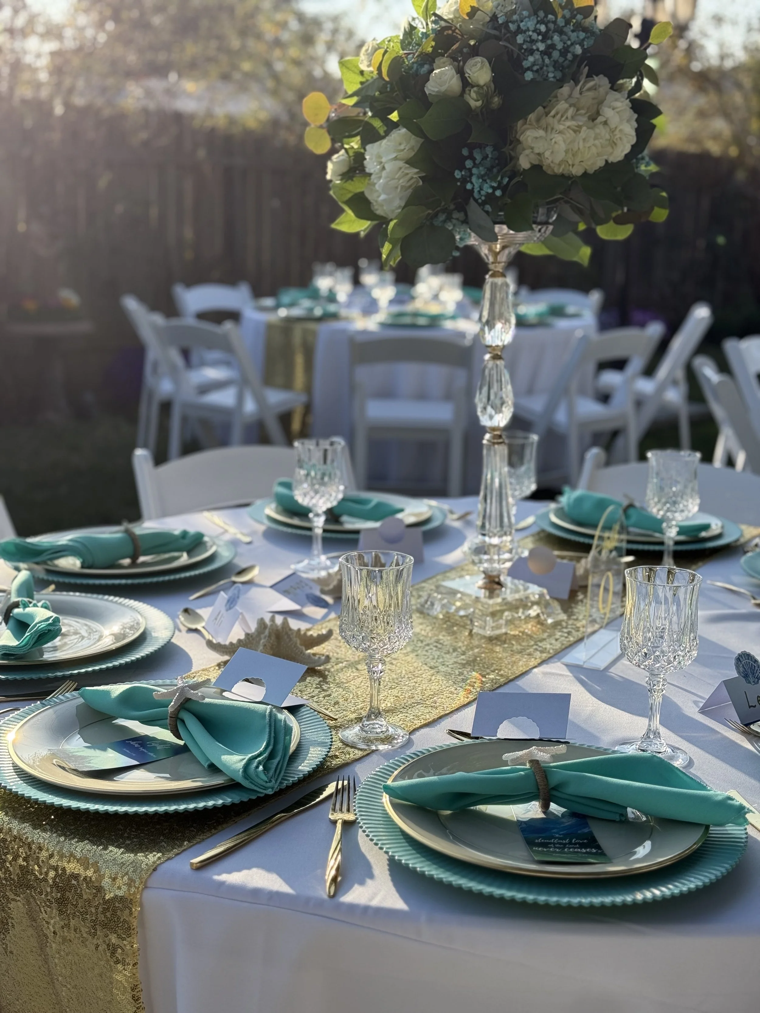 Elegant outdoor table setting with white tablecloths, teal napkins, gold cutlery, crystal glassware, and a floral centerpiece.