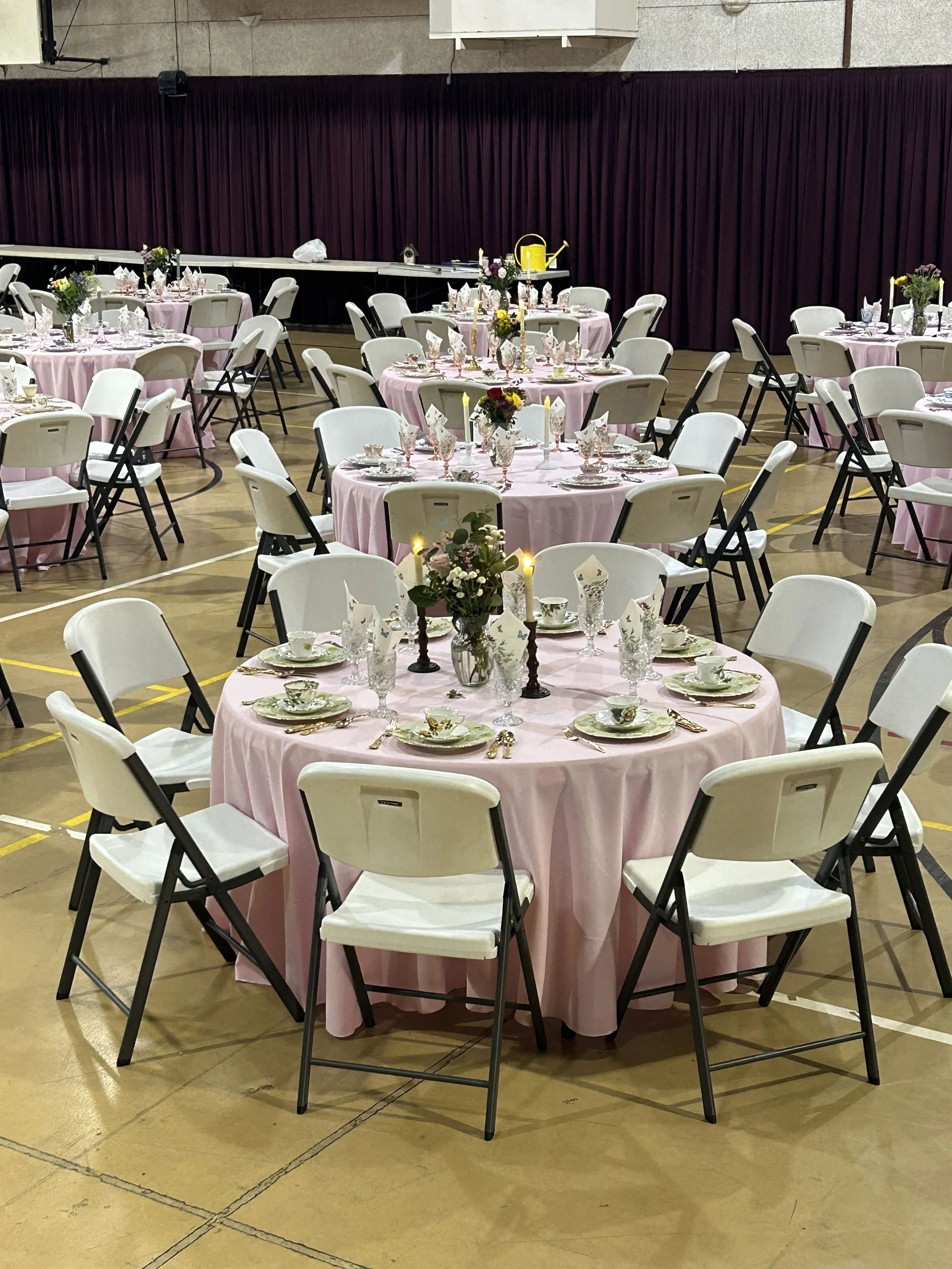 Indoor event setup with round tables covered in pink tablecloths, white folding chairs, and elegant table settings with floral arrangements and candles.